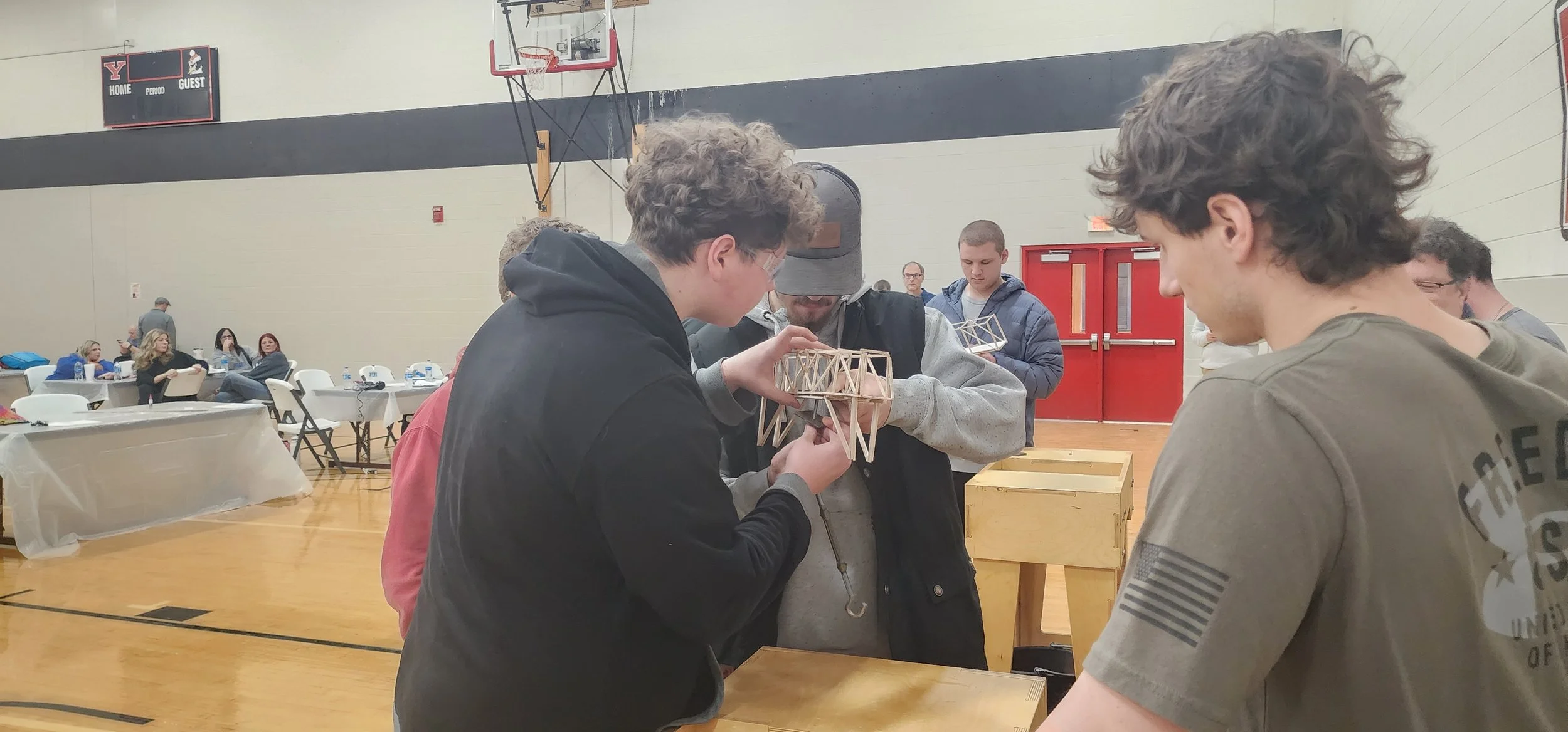 Group of young men examining a small wooden bridge model in a gymnasium, with tables and people in the background.