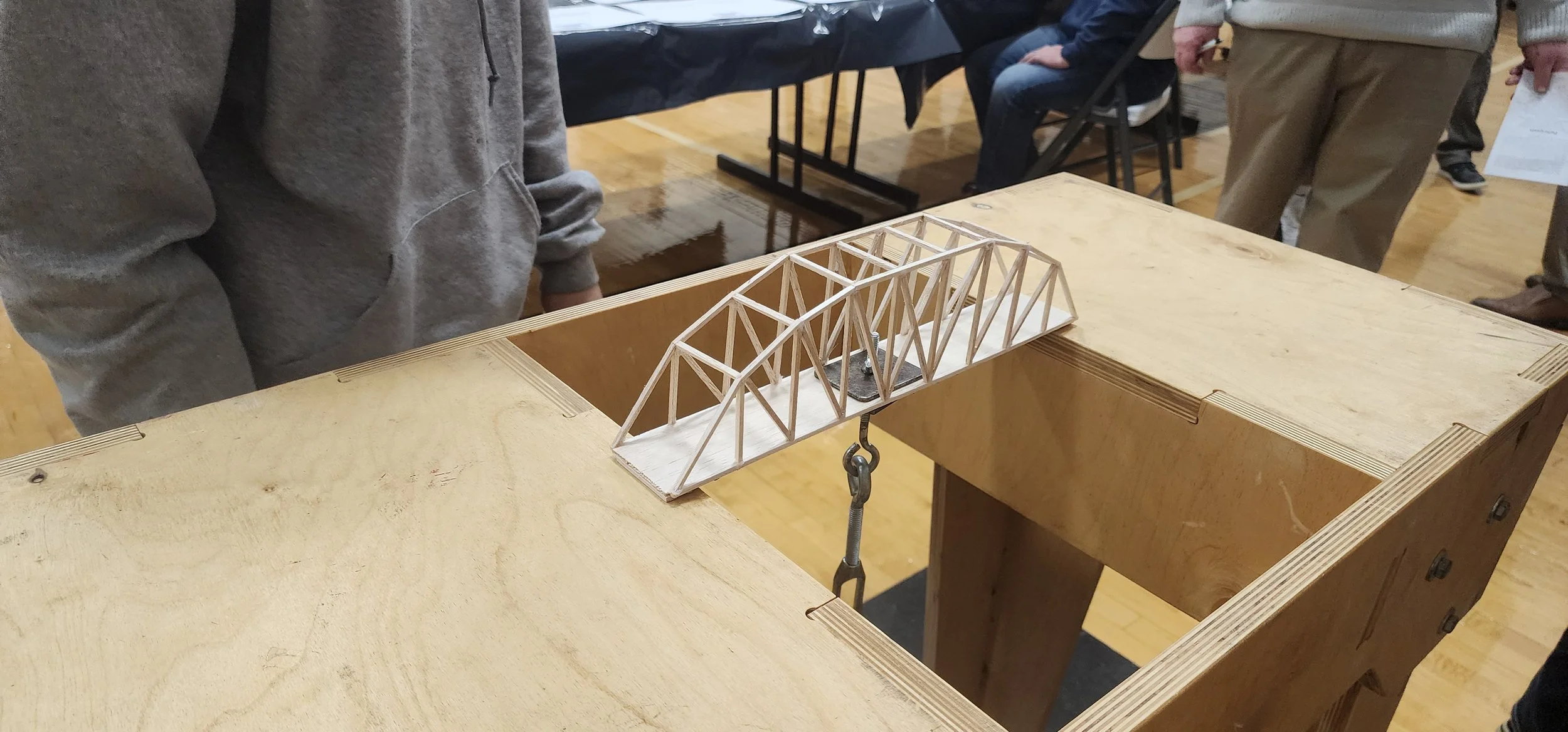 A miniature wooden bridge model displayed on a wooden table at an indoor event, with people observing.