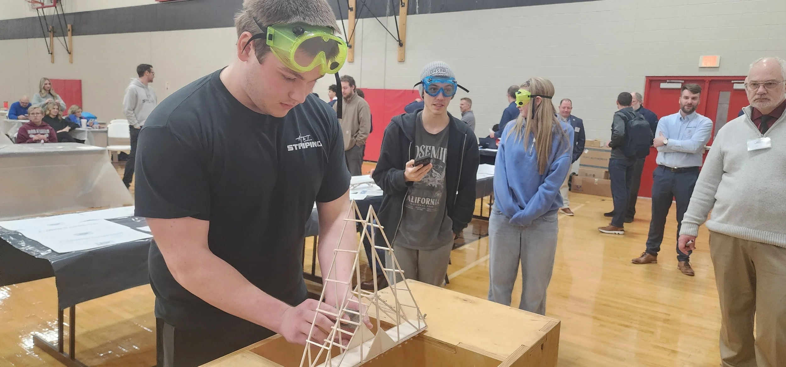 Young man wearing safety goggles builds a wooden structure at a table during an indoor event, with people and exhibitors observing.