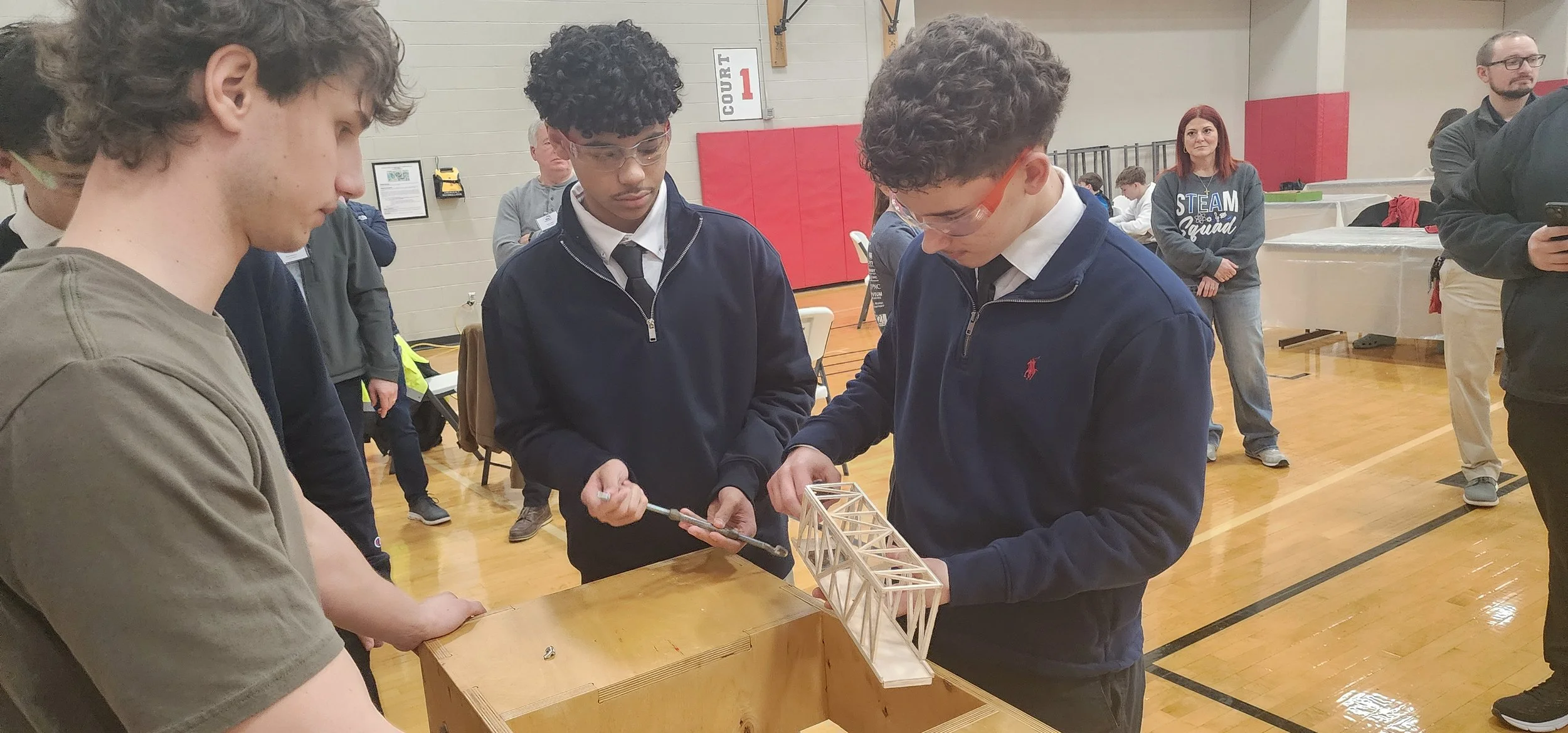 Group of young students working together on a wooden bridge project at an indoor event.