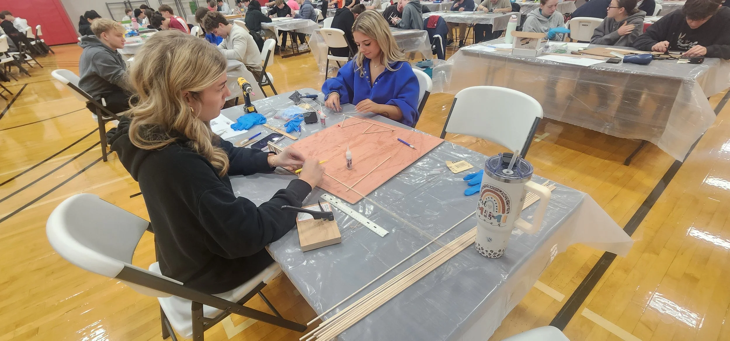 Two young women working on a wood project at a table in a crowded indoor workshop or event. They have tools, sticks, and glue on the table. The room has many other people working at tables.