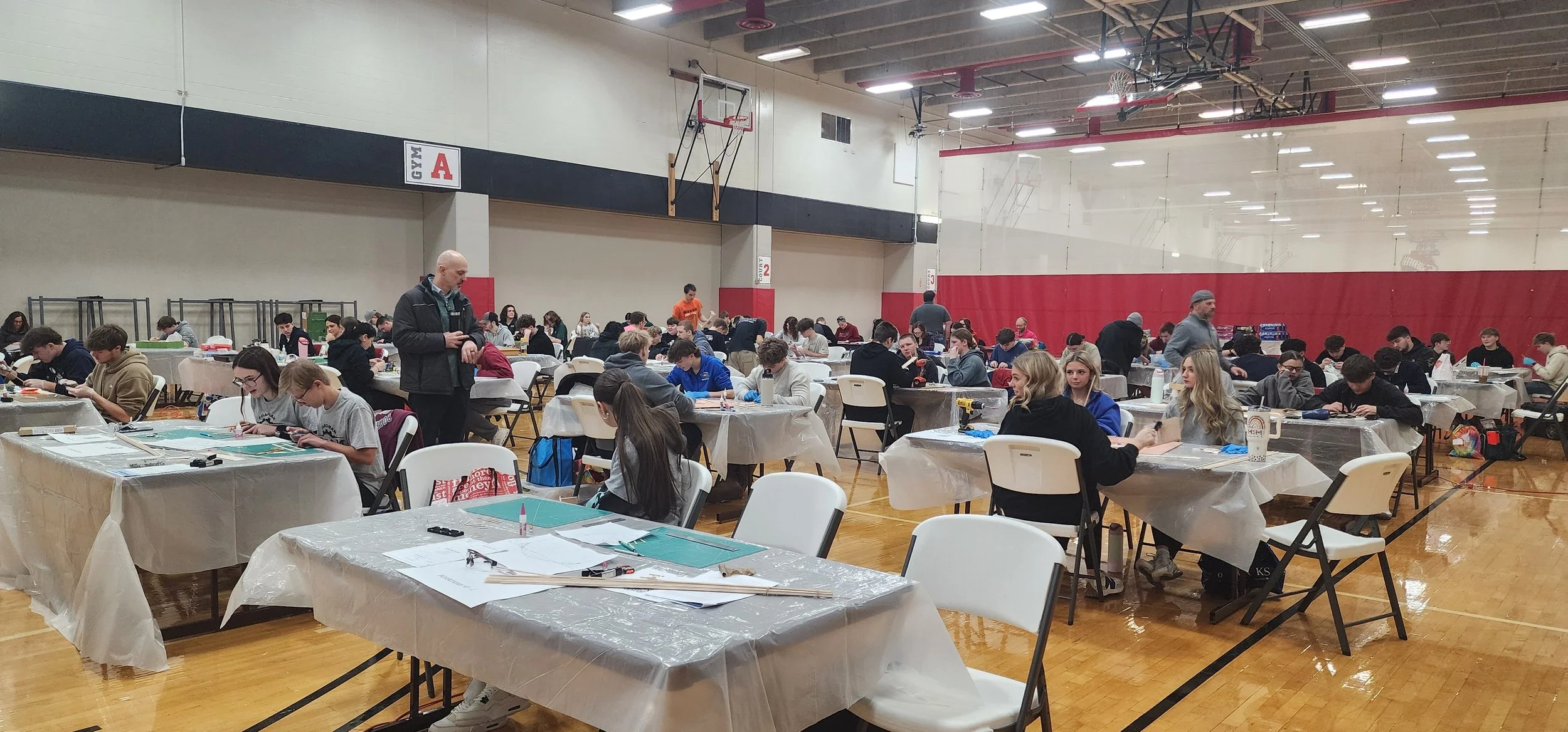 A large indoor gymnasium filled with students sitting at tables covered with plastic tablecloths, working on projects. There are adults standing and supervising. The gym has a basketball hoop and wood flooring, with red and black walls.