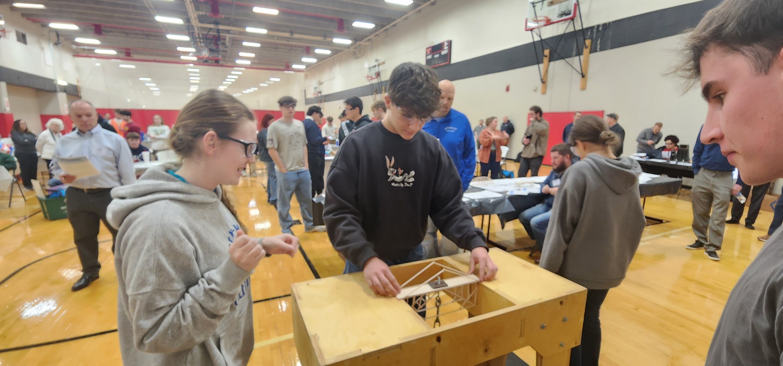 Young people playing a tabletop game at a busy indoor event inside a gymnasium, with many attendees and tables in the background.