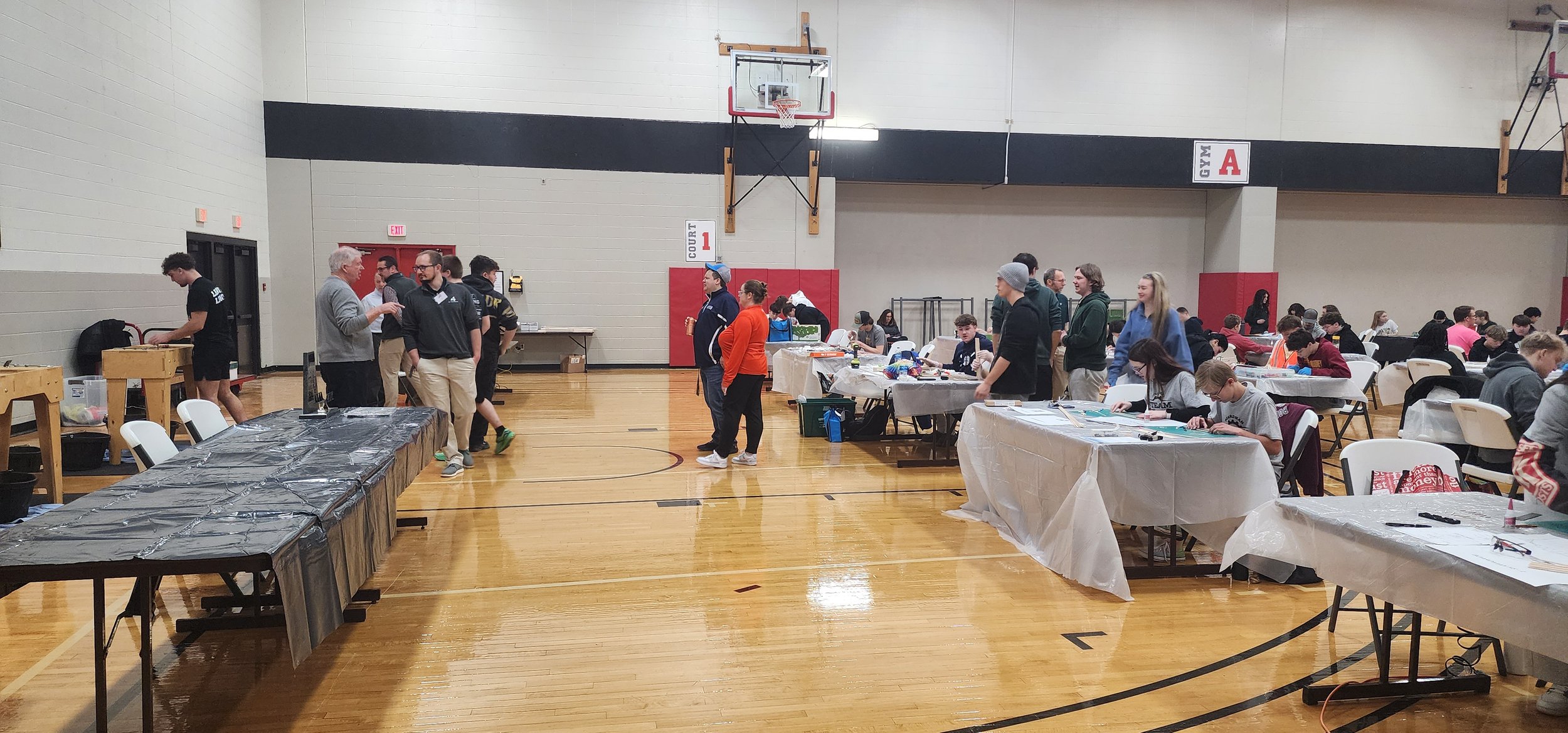 A gymnasium filled with people working at tables and displays, with some standing and chatting, children sitting and working, and tables covered with various items and supplies.