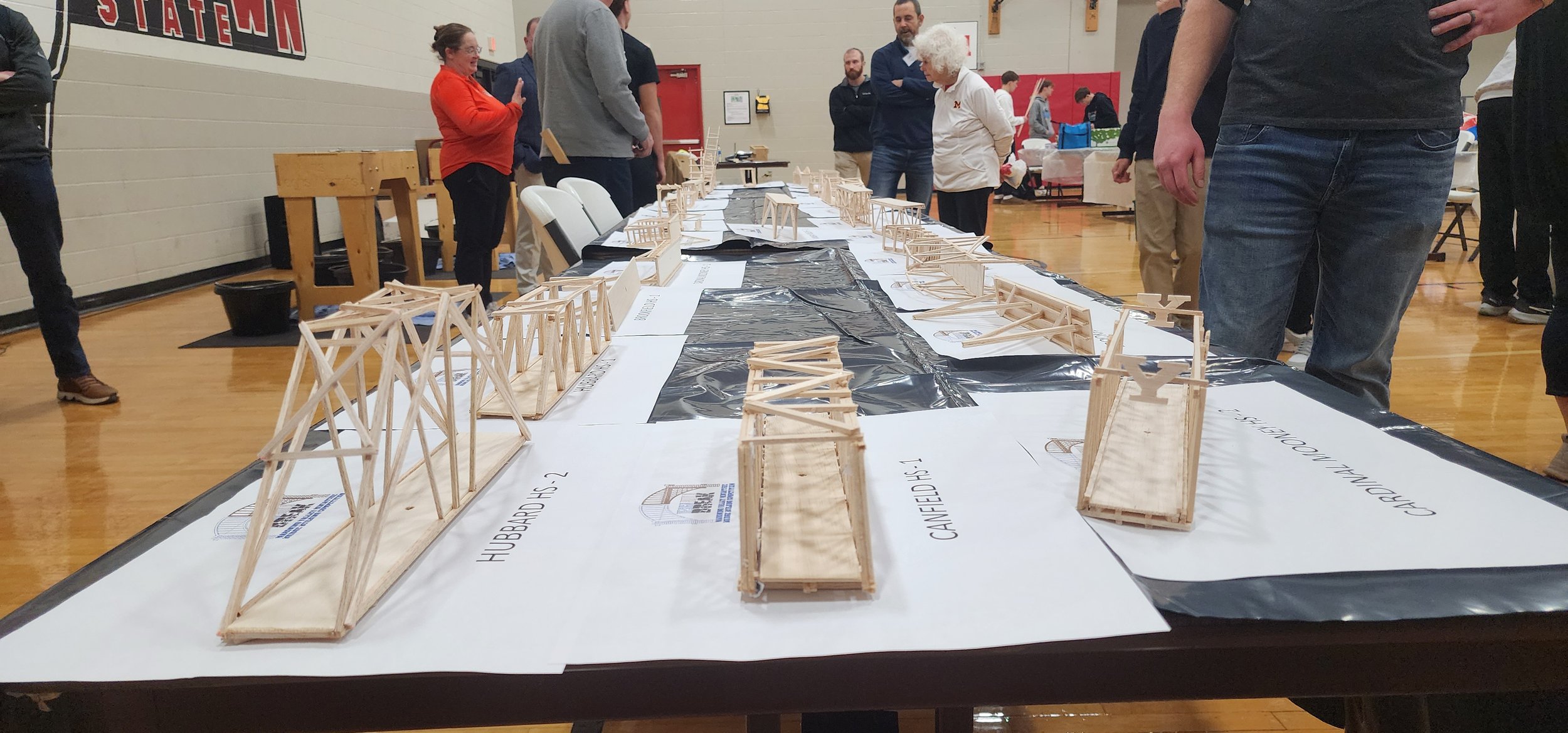 People gathered around tables displaying various miniature bridge models and architectural structural designs in a gymnasium.