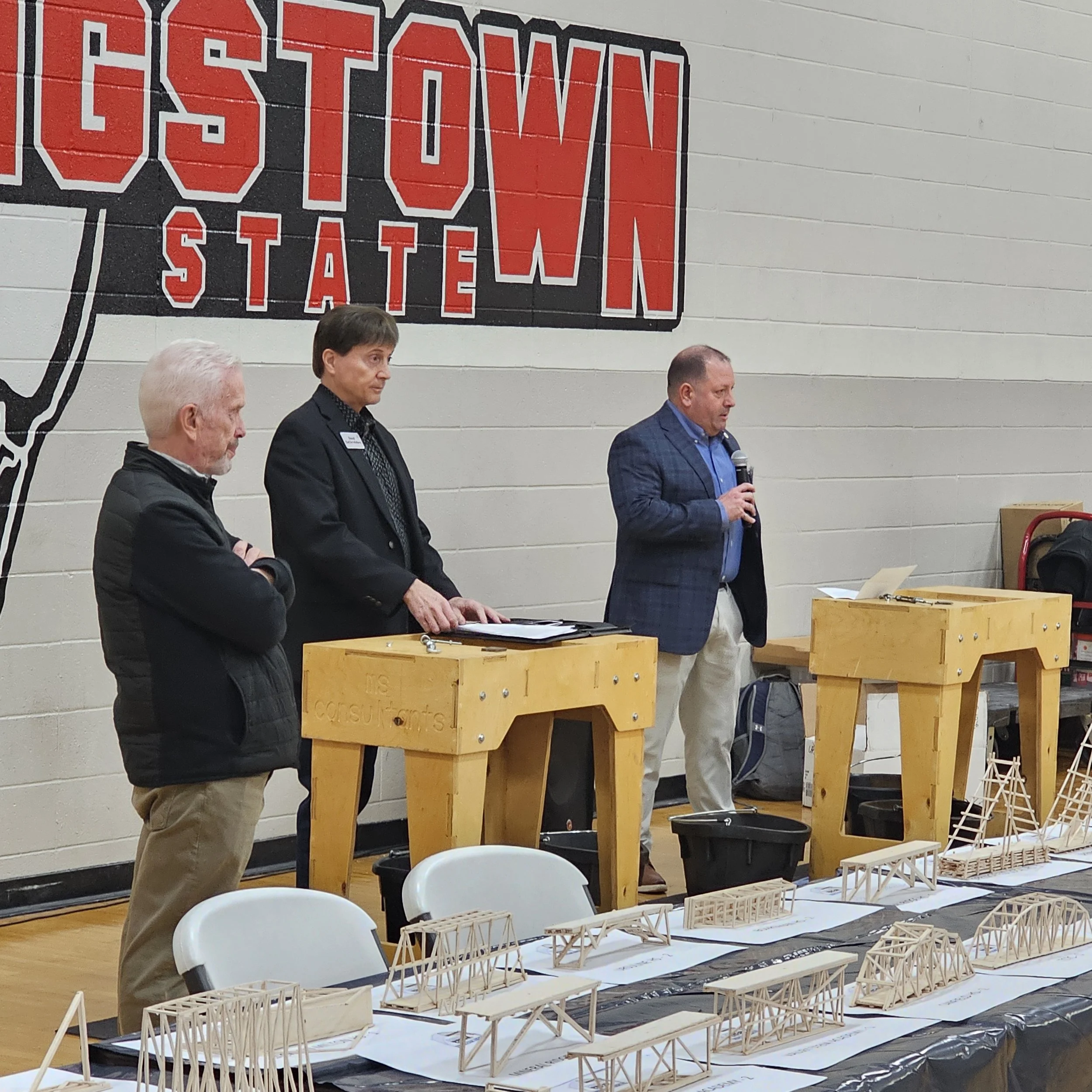 People standing behind tables with architectural models displayed at an event, in front of a large Mississippi State sign on a brick wall.