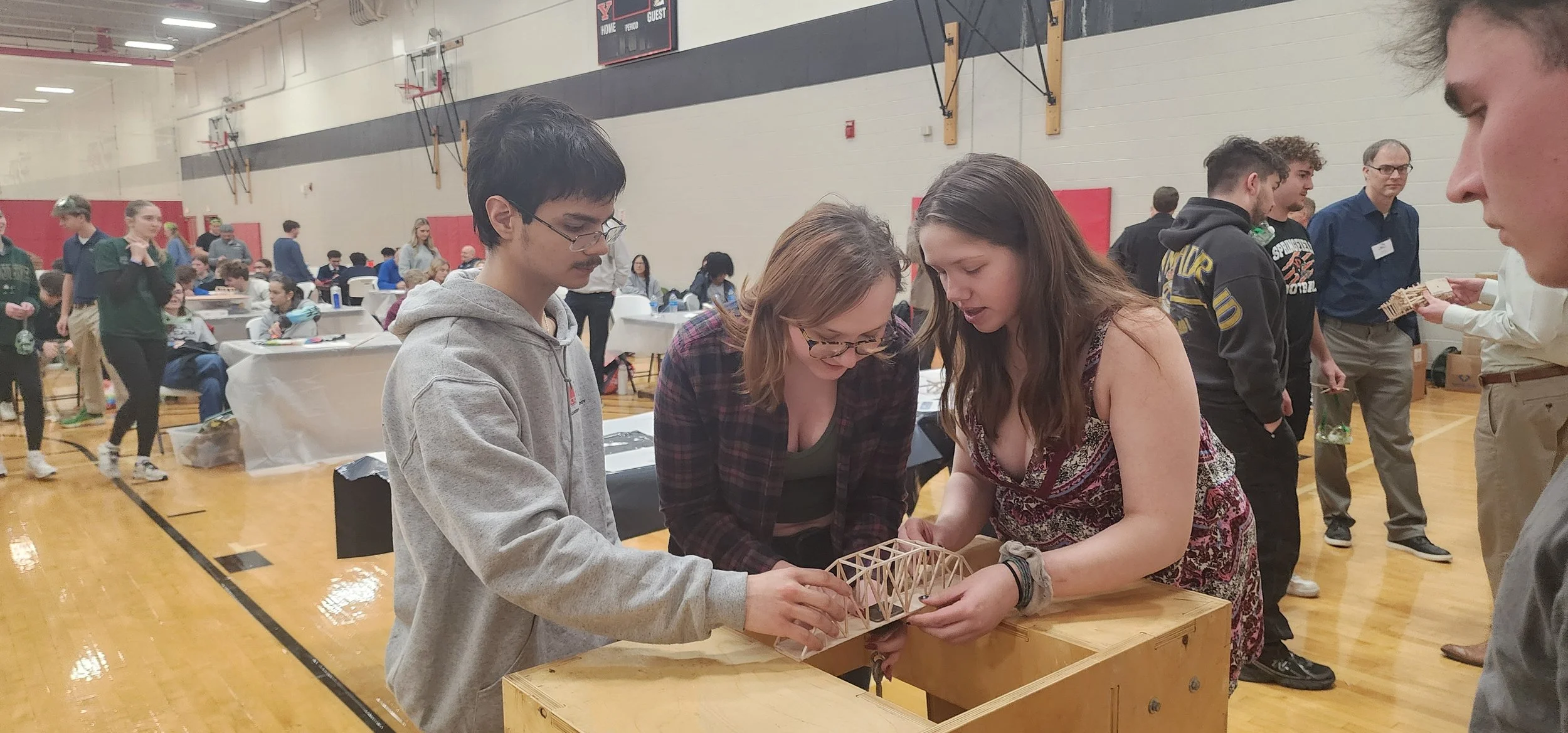 Group of people gathered around a table in a gymnasium, examining a wooden bridge model, with others in the background