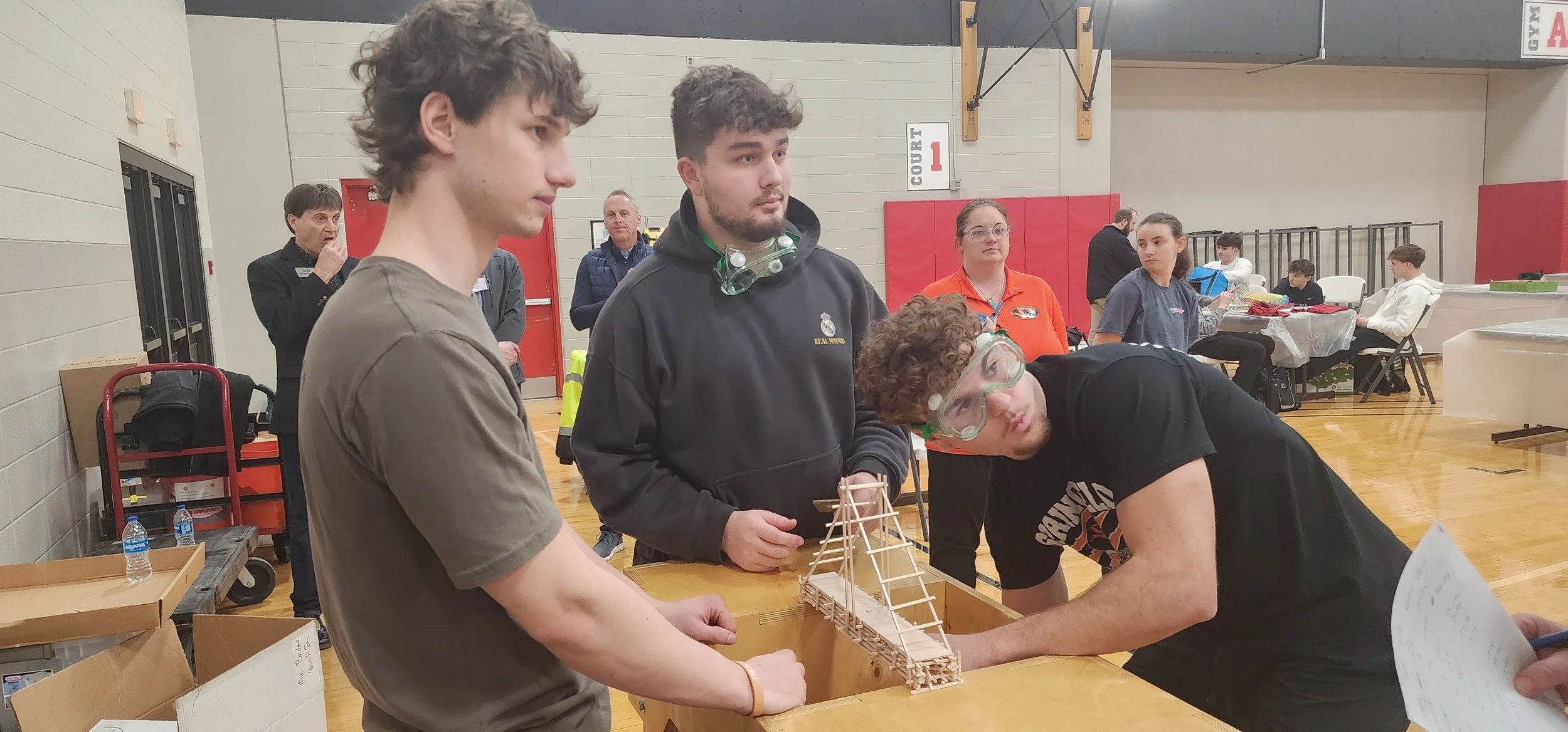 Group of young people working on a bridge model in a gymnasium, some wearing goggles, with a few adults observing in the background.