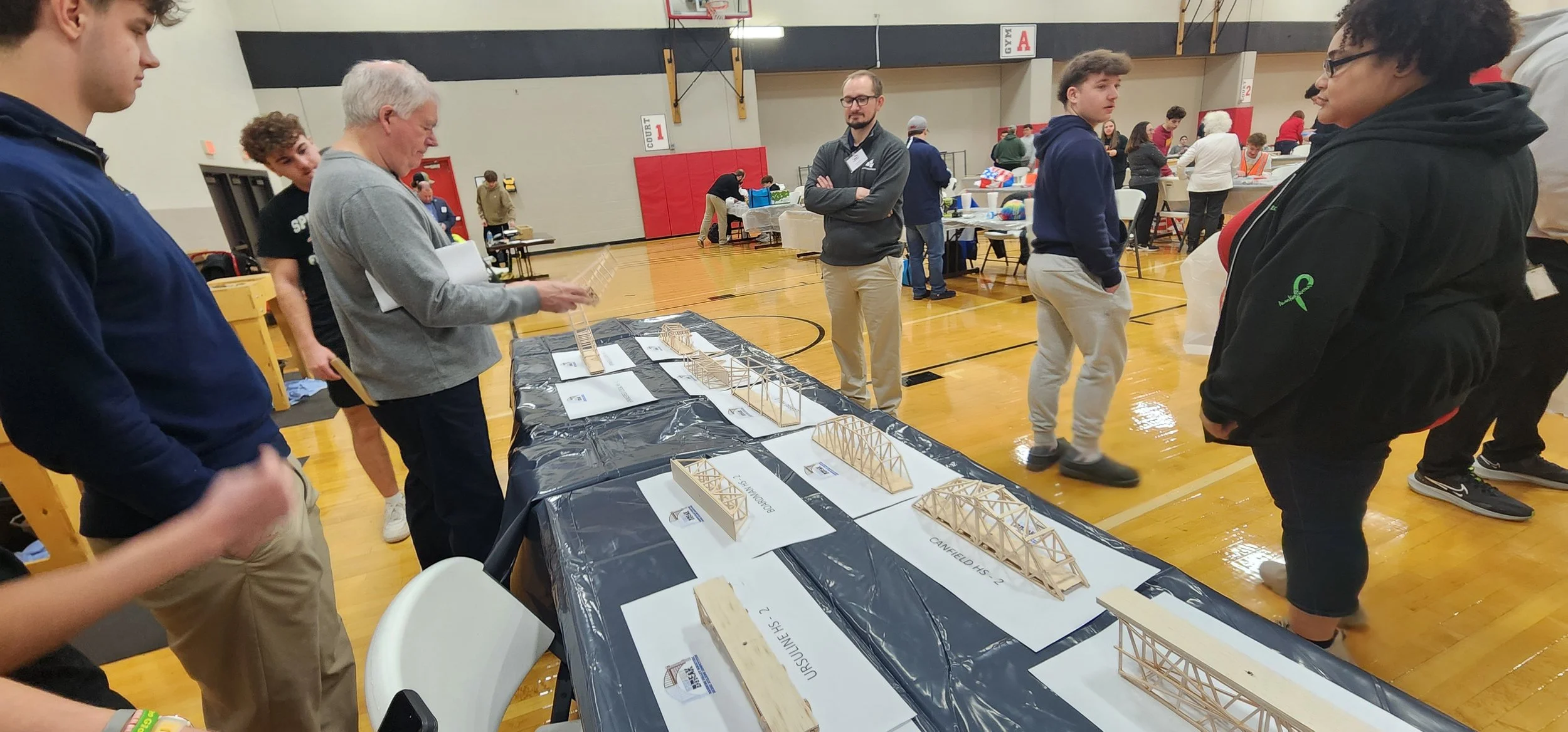 A group of people gathered around a table displaying small wooden bridge models, with some individuals closely examining the models and others engaging in conversation, inside a gymnasium.