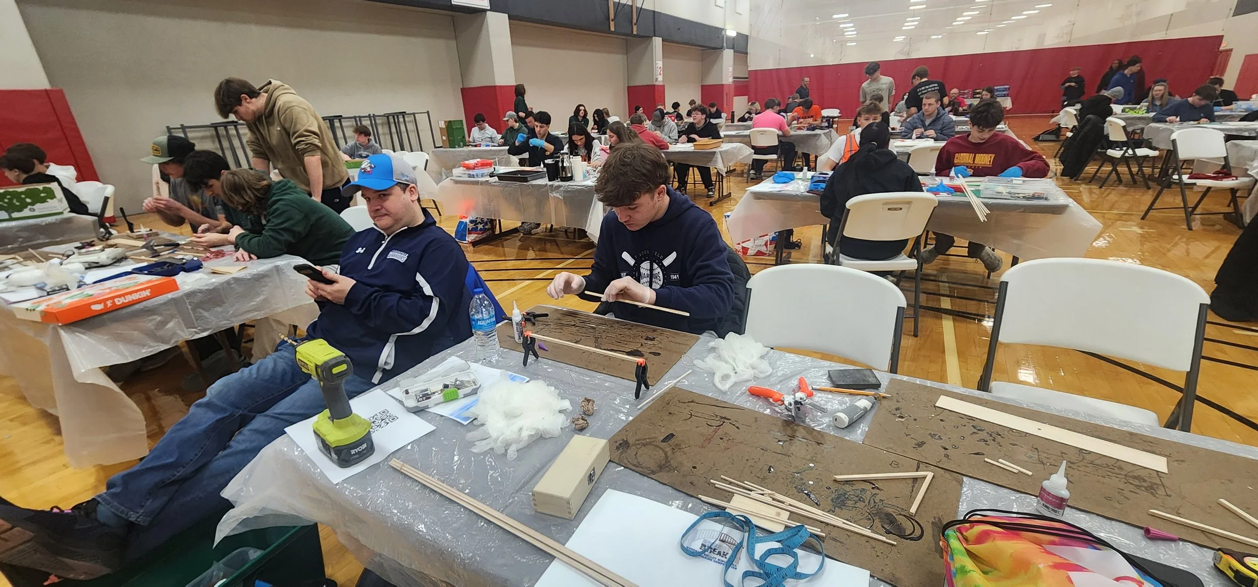 A large indoor space filled with many people sitting at tables and working on projects, with various materials and tools scattered across the tables. Some people are wearing gloves, and the atmosphere suggests a workshop or crafting event.