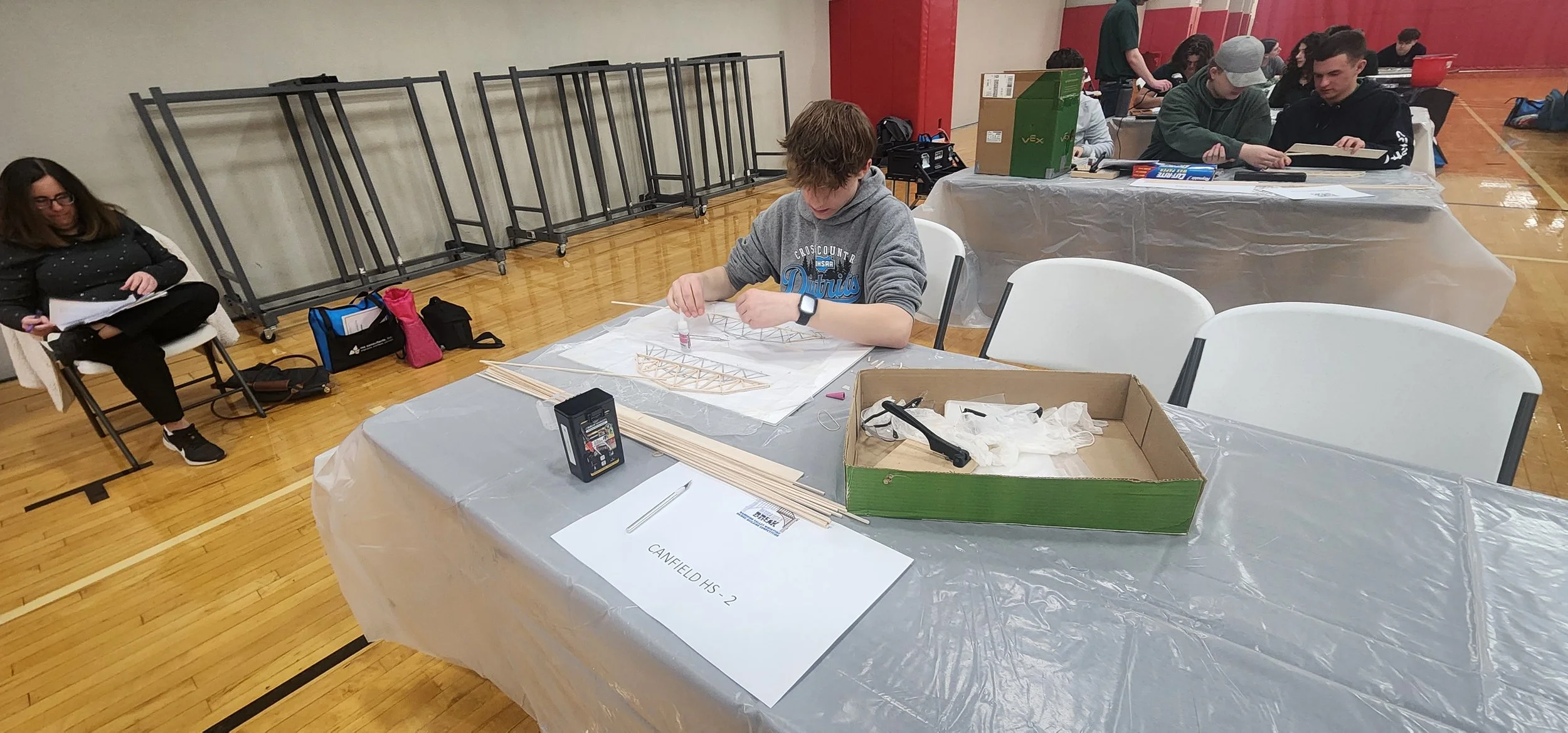 Students working on architecture or engineering projects at tables in a school gymnasium during a workshop or competition.
