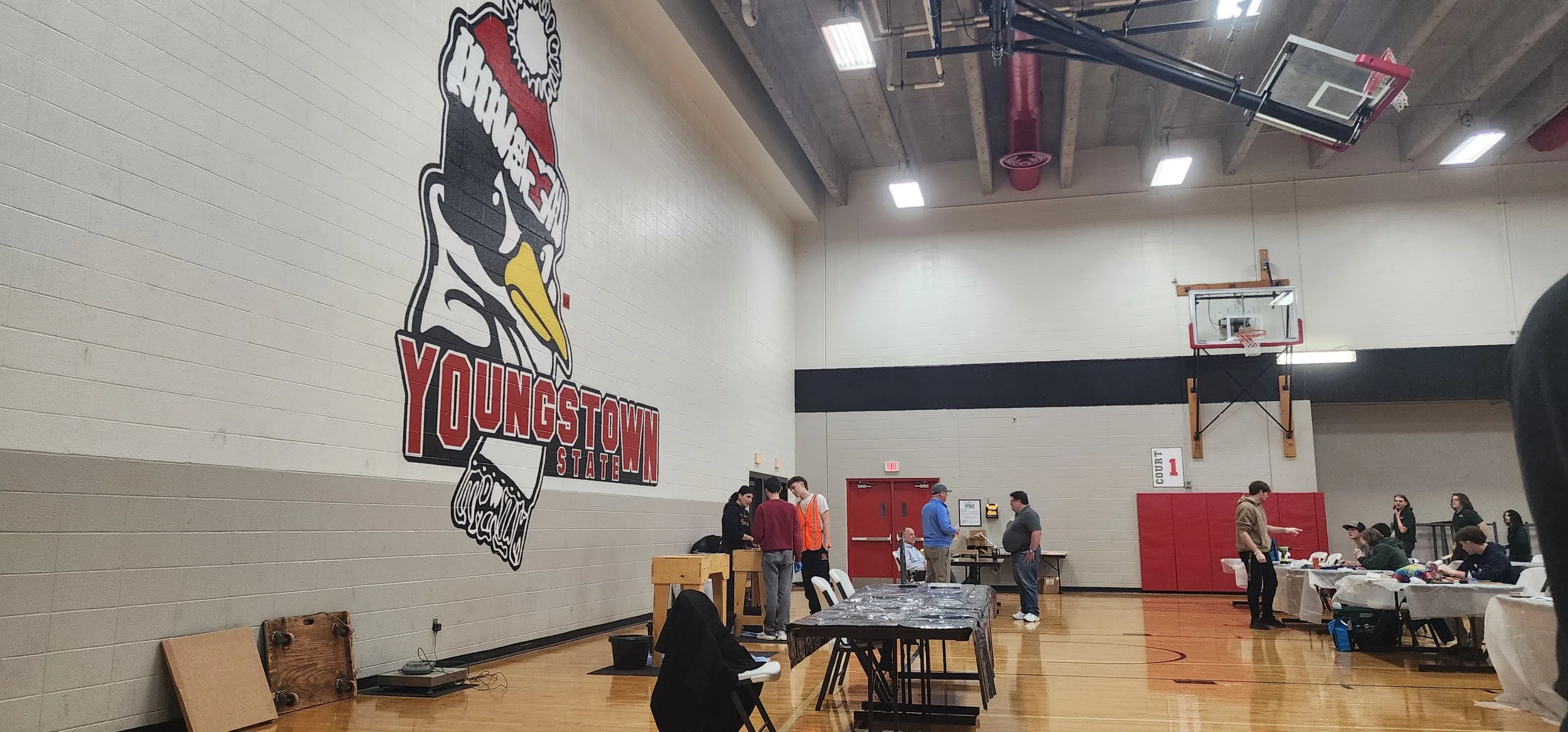 Indoor gymnasium with a large Youngstown State logo on the wall, several people standing and talking near tables, a basketball hoop on the far wall, and tables with items on them.
