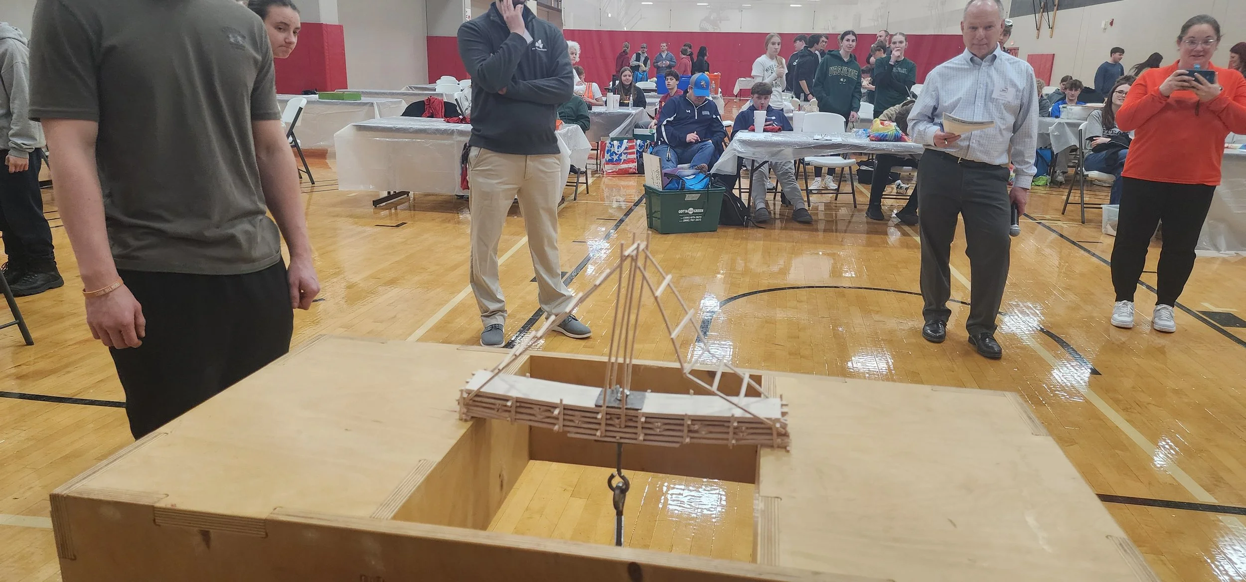 People gathered in a gymnasium, observing a small wooden bridge bridge model on a table in the foreground.