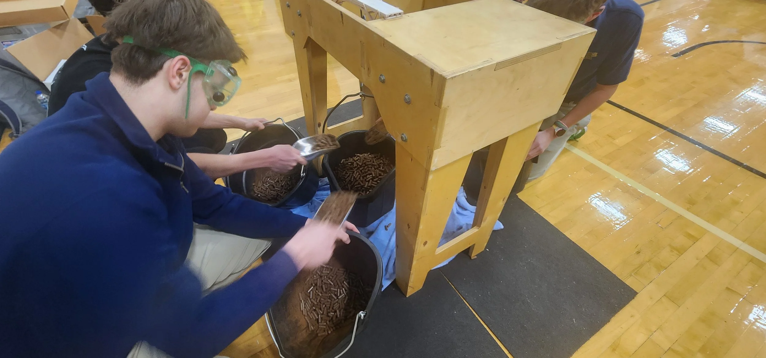 A person in safety goggles scooping chocolate sprinkles from a bucket into a bin in a gymnasium.