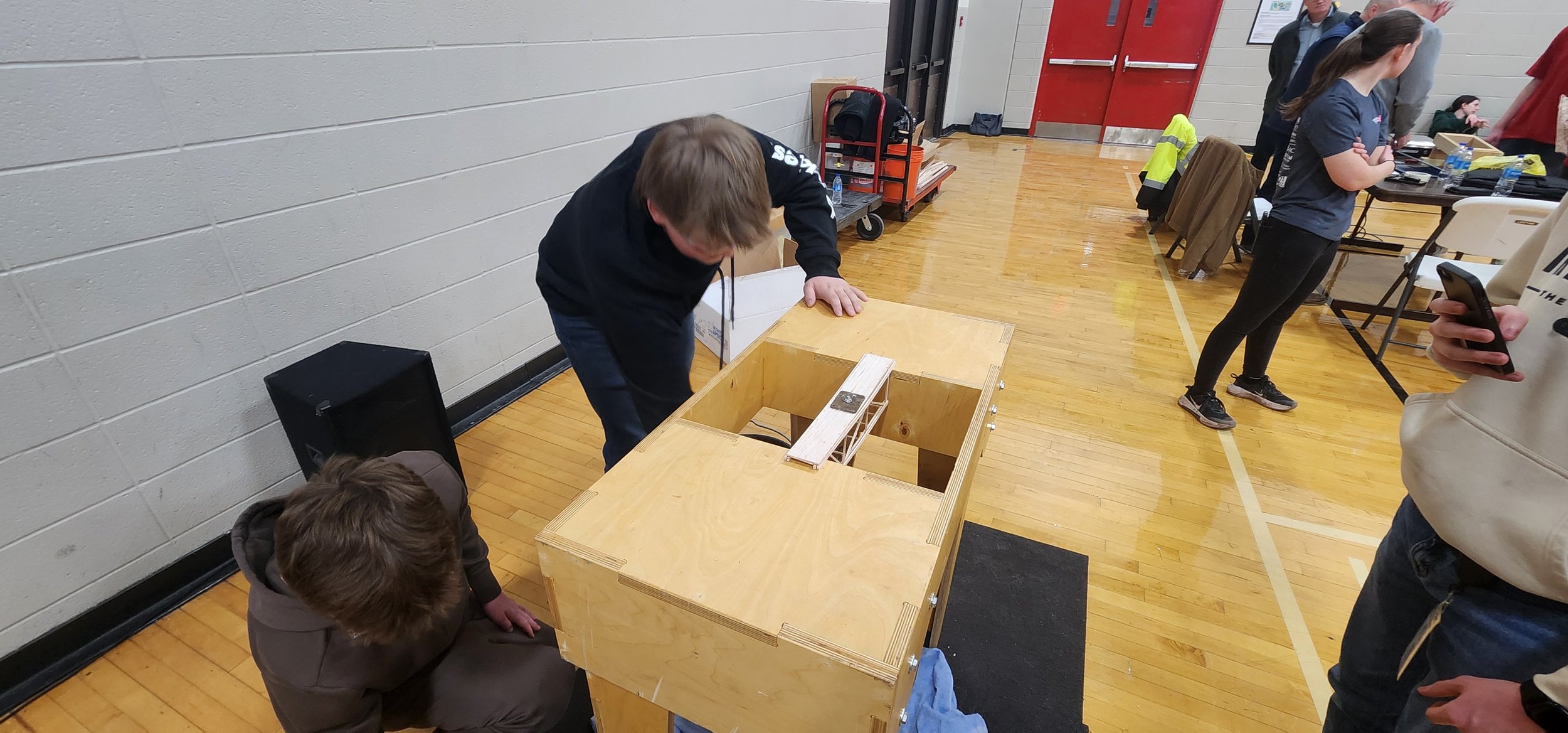 A group of people gathered around a wooden voting box in a gymnasium, with some people casting ballots and others waiting or observing.