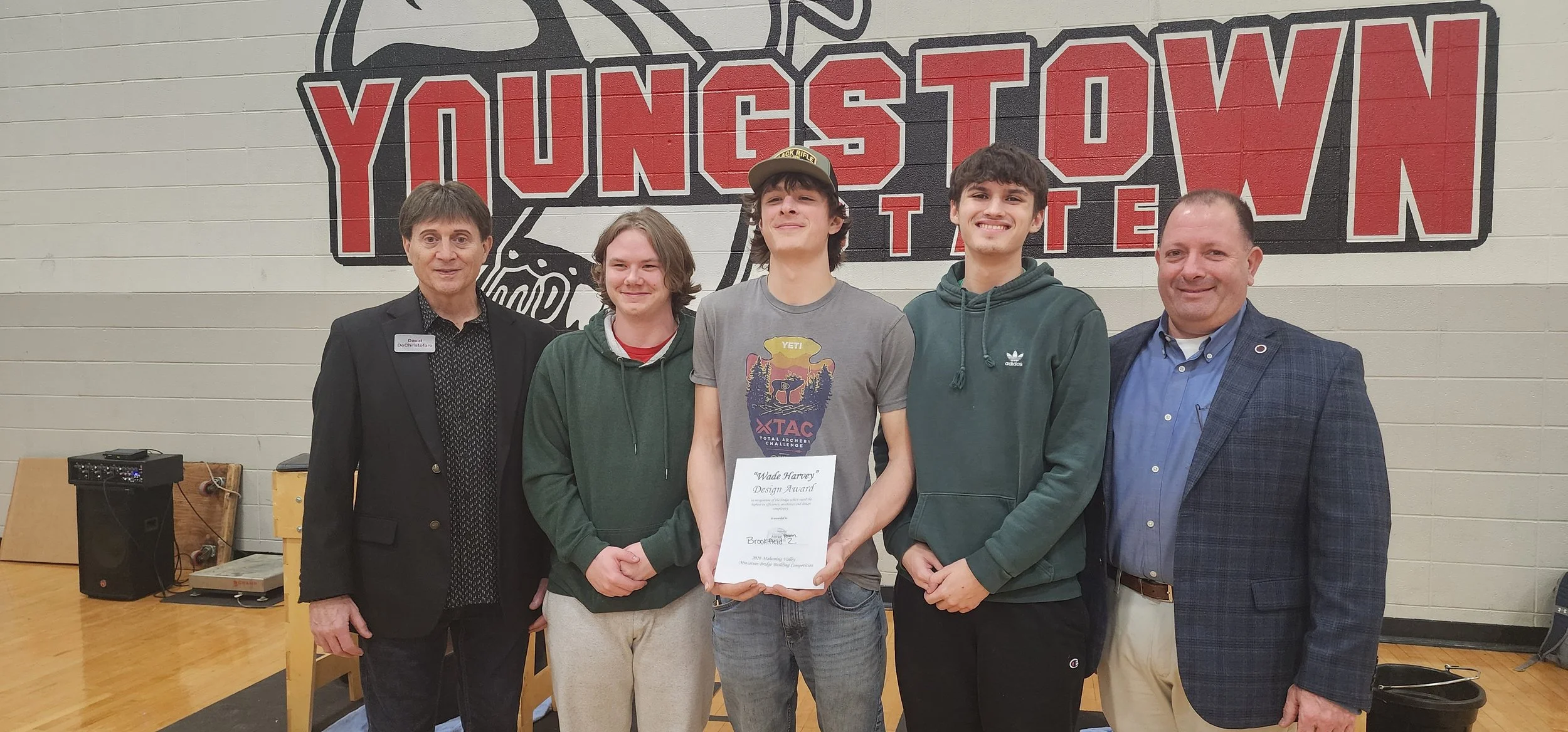 A group of five males standing in front of a wall with a large logo that reads 'Youngstown State'. The person in the center is holding a certificate, and there are two men on each side of him. They are in an indoor gym or event space, with equipment 