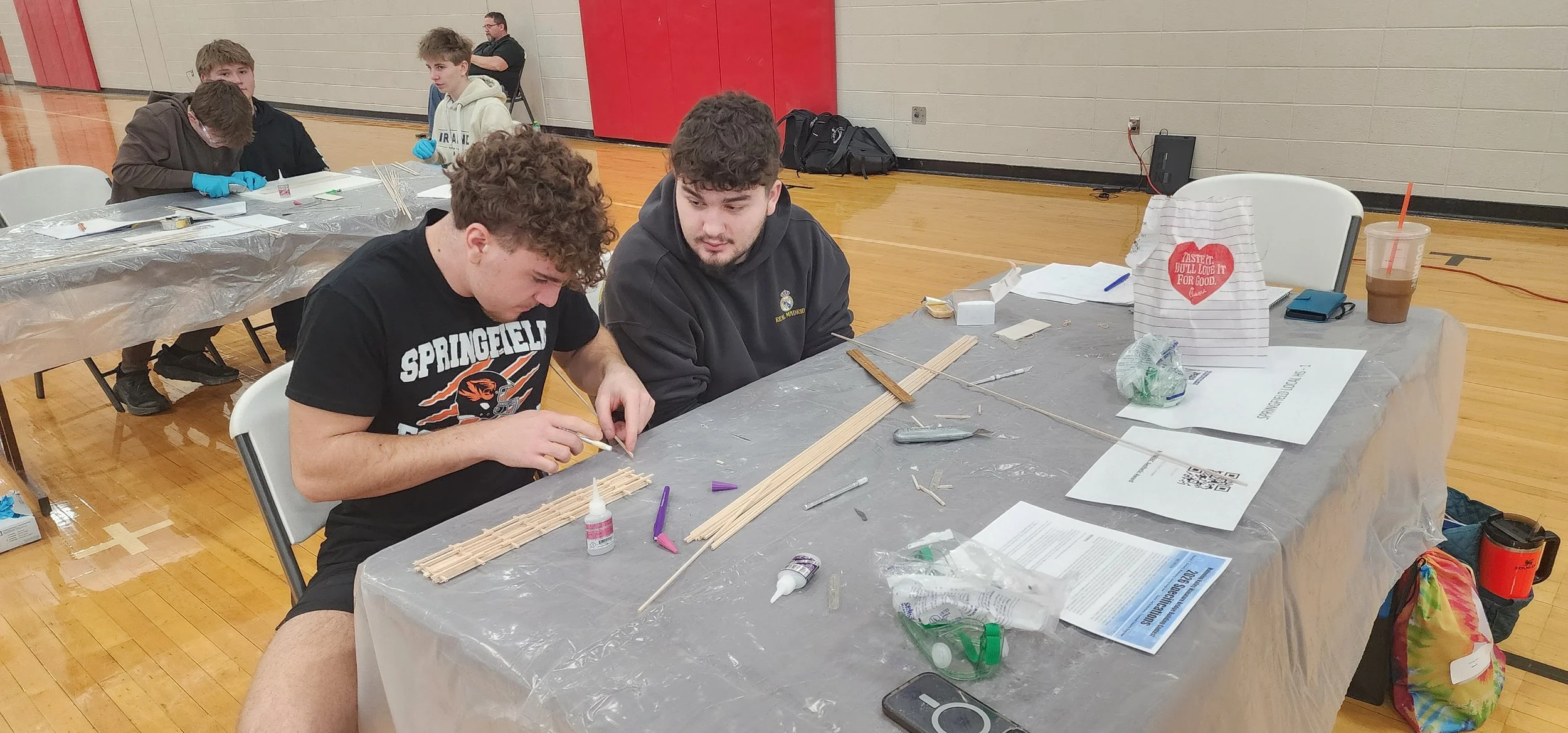 Two young men sit at a table working on a woodworking project with wooden sticks, glue, and tools, in a gymnasium or indoor sports hall, with other young people in the background.