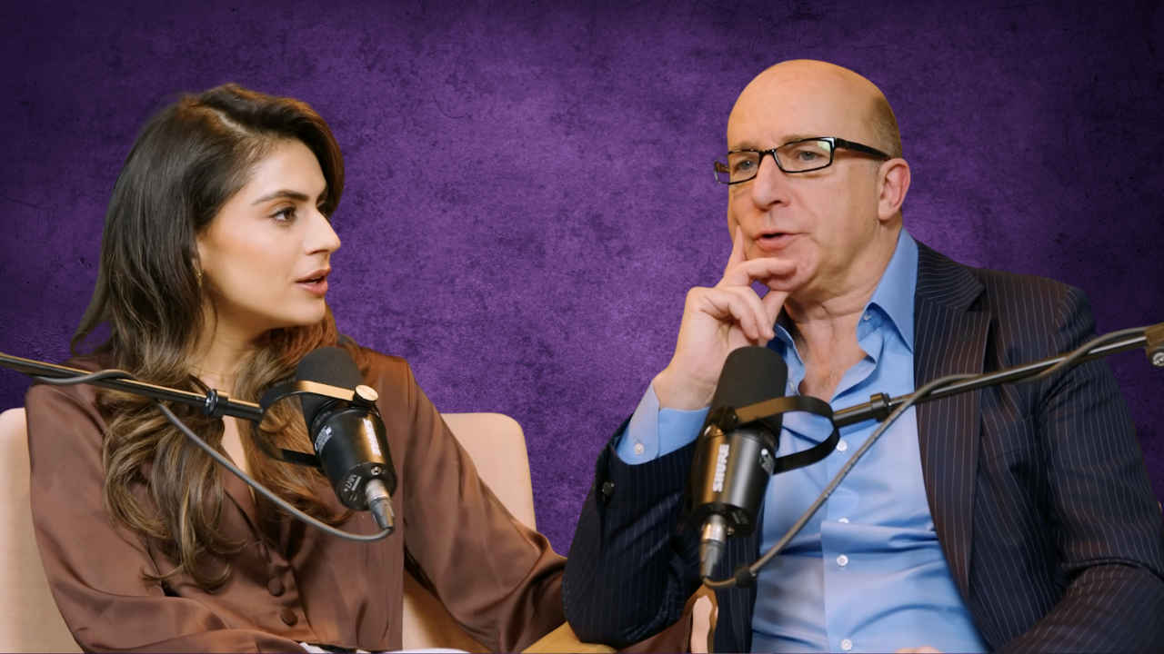 A woman and a man sit at a table with microphones, engaged in conversation. The woman has long brown hair, wearing a brown silky blouse, and is looking at the man. The man is bald, wears glasses, a dark pinstripe blazer over a light blue shirt, and is resting his chin on his hand while listening.