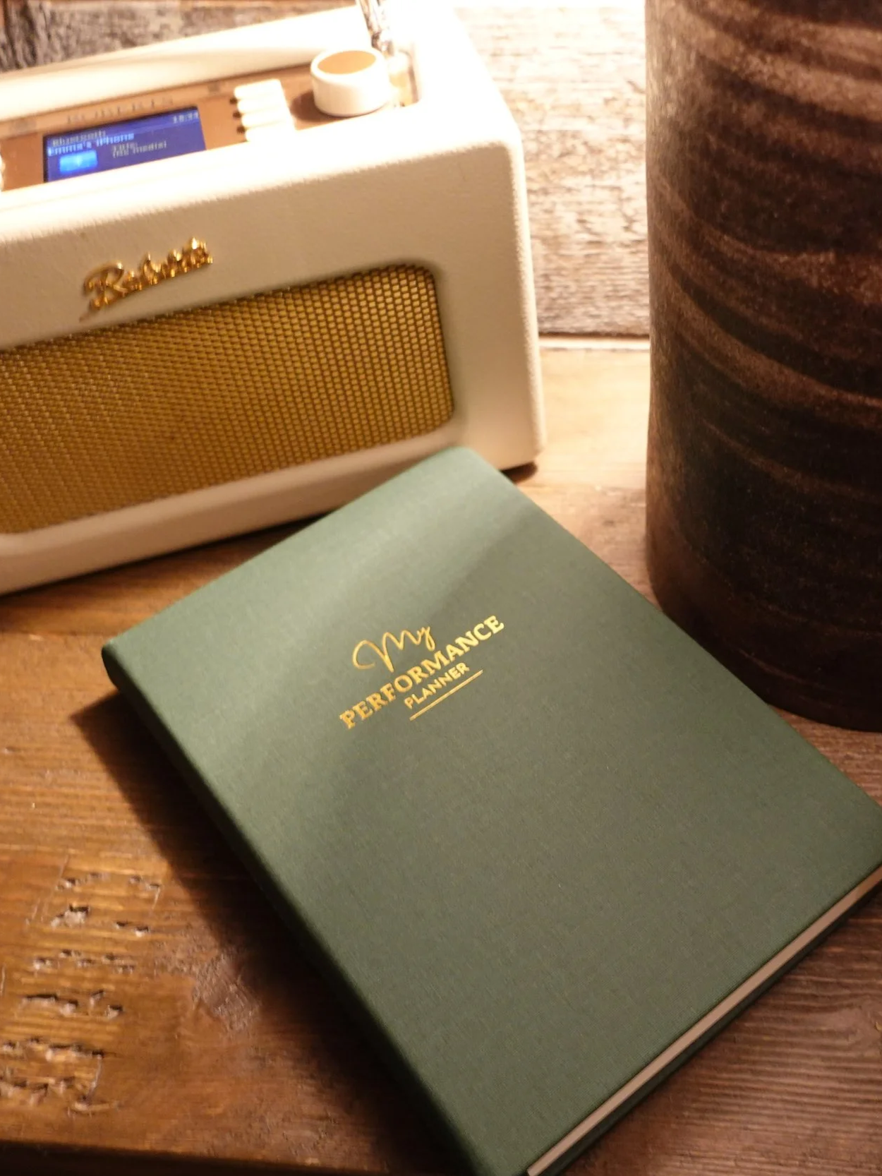 A desk scene with a green performance planner, a white radio, and a dark textured vase on a wooden surface.