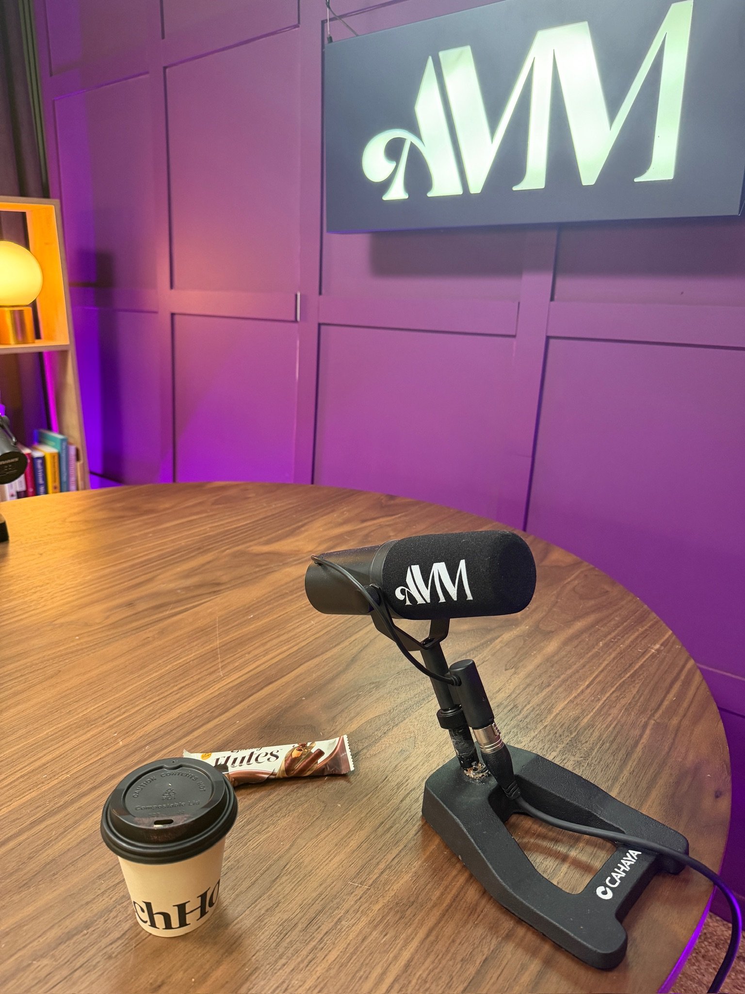 A round wooden table with a black microphone on a stand, a paper coffee cup with a black lid, a chocolate snack bar, and a camera lens cap, with a purple wall and a large illuminated sign in the background.