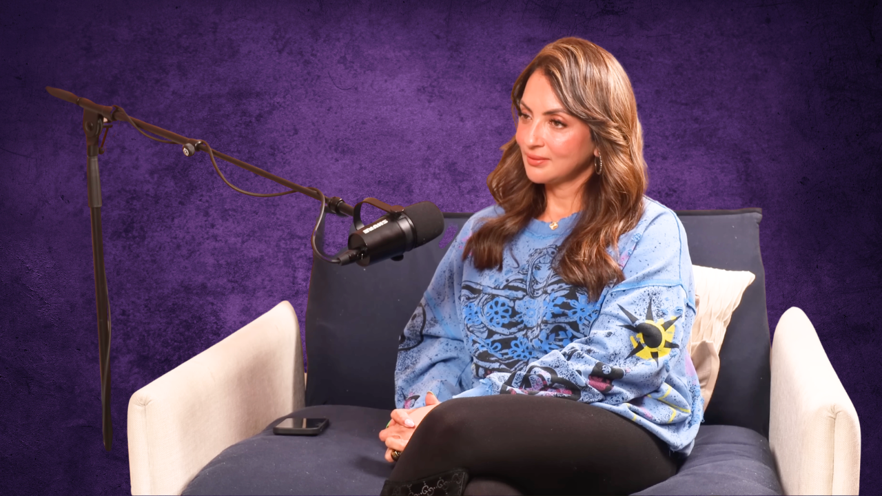 Woman with long brown hair sitting on a couch with a purple textured background, holding her hands on her lap, with a microphone positioned in front of her.