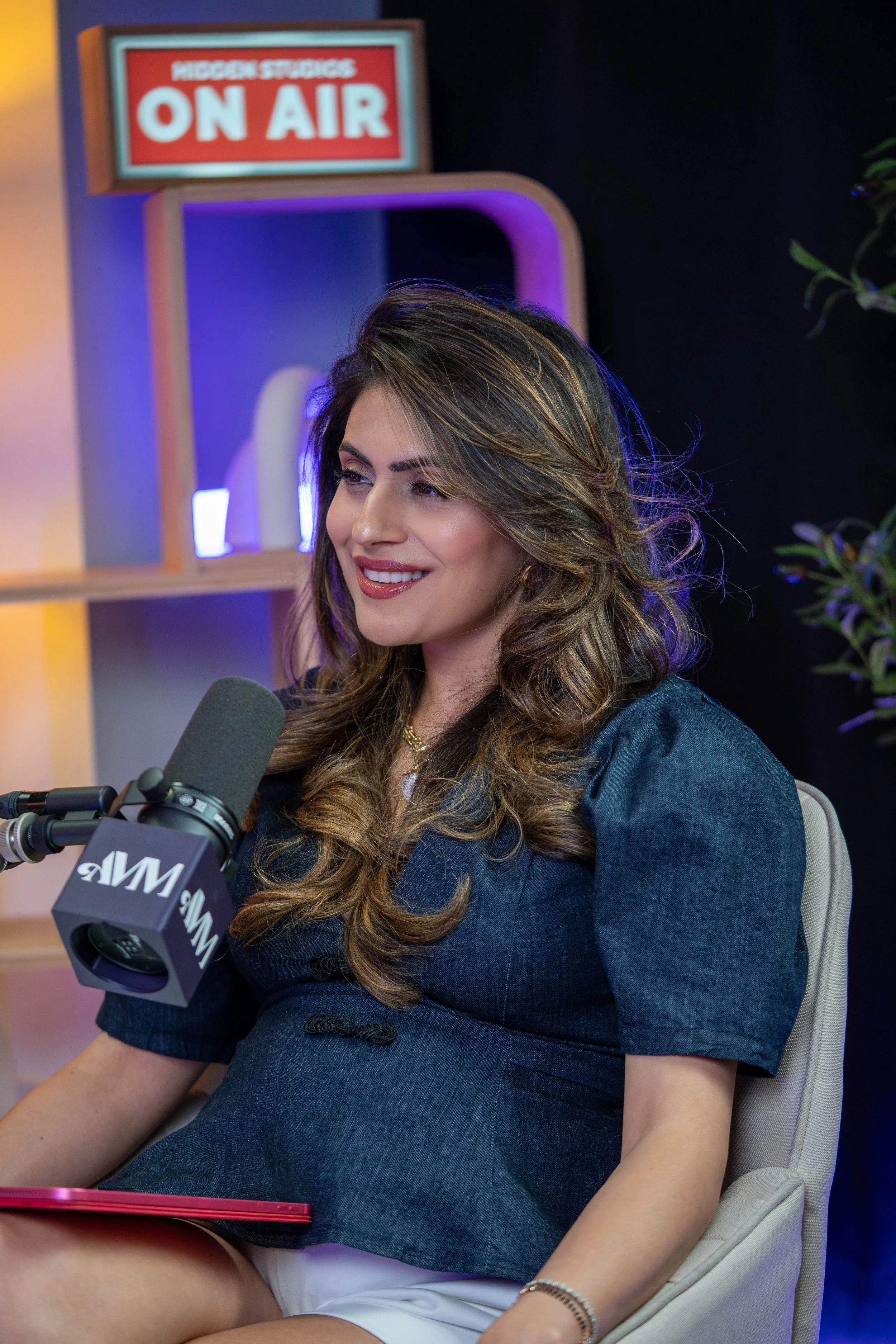A woman with long wavy hair, smiling, is sitting in a chair with a pink tablet on her lap. She is speaking into a microphone labeled 'MSNBC.' Behind her, there is a sign that says 'MOGGAN STUFFS ON AIR' and a decorative shelf with purple and yellow lighting, along with a plant on the right side.