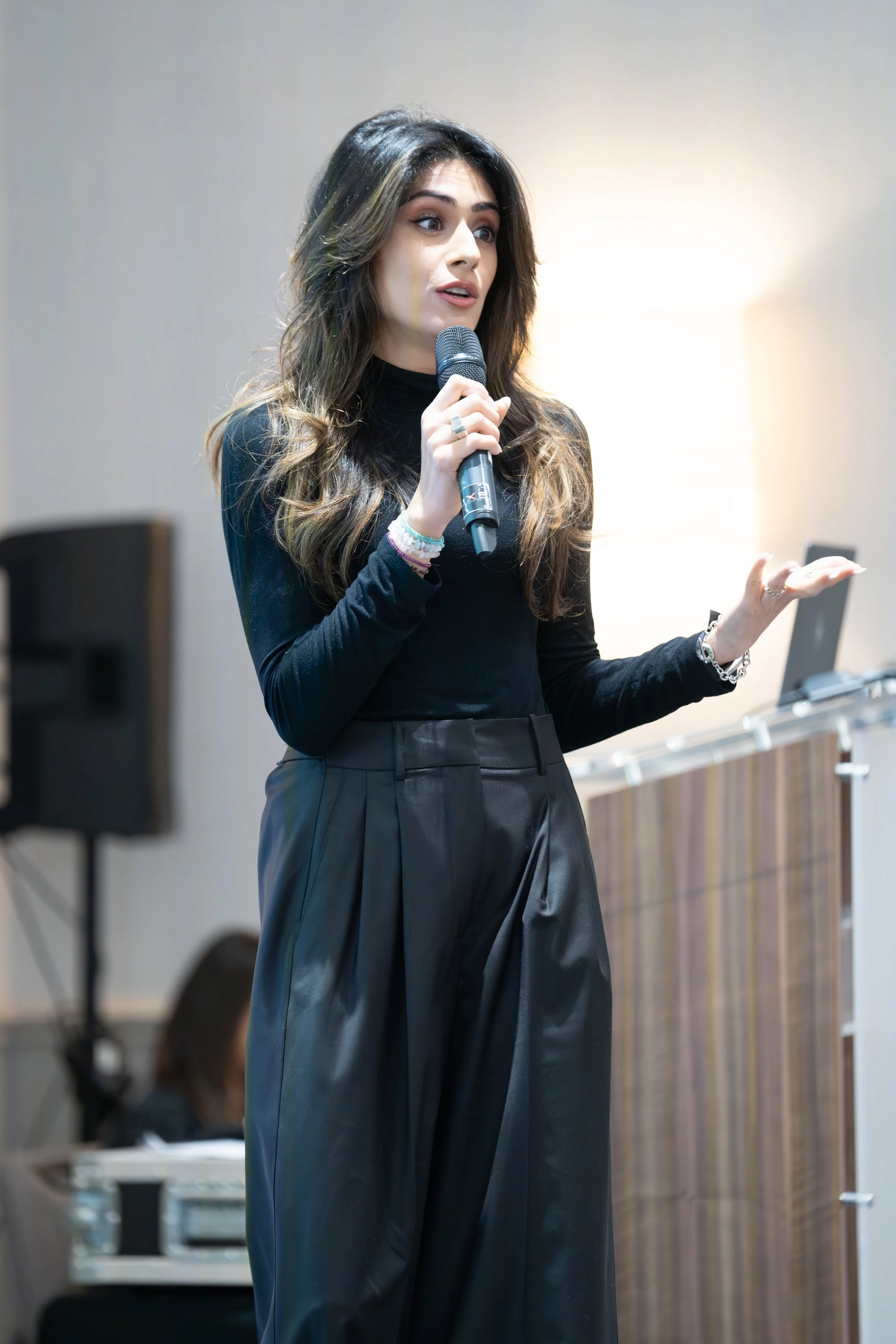 A young woman with long, wavy brown hair, speaking into a microphone during a presentation or speech. She is wearing a black long-sleeve top and black high-waisted pants.