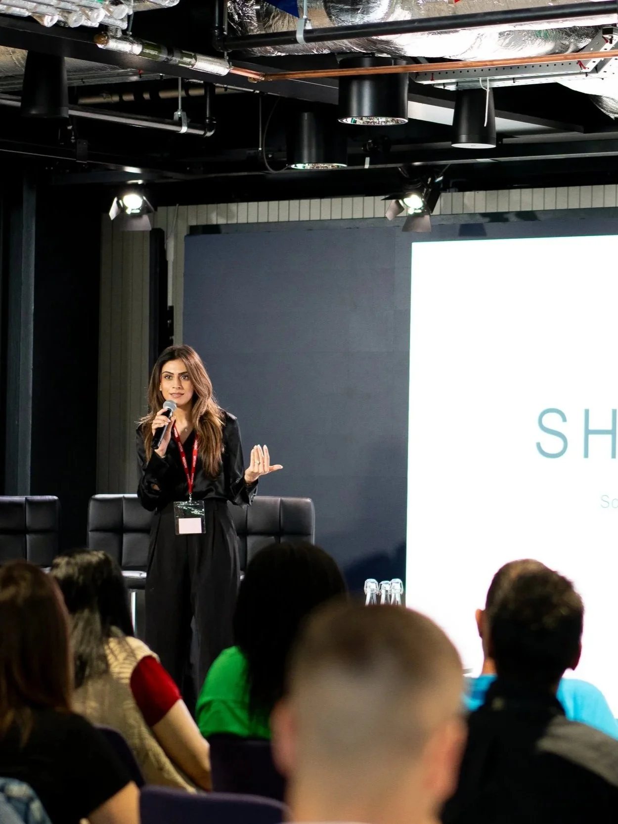 A woman speaking into a microphone during a conference presentation. She is standing in front of a large screen with part of a word visible. The audience is seated, facing her.