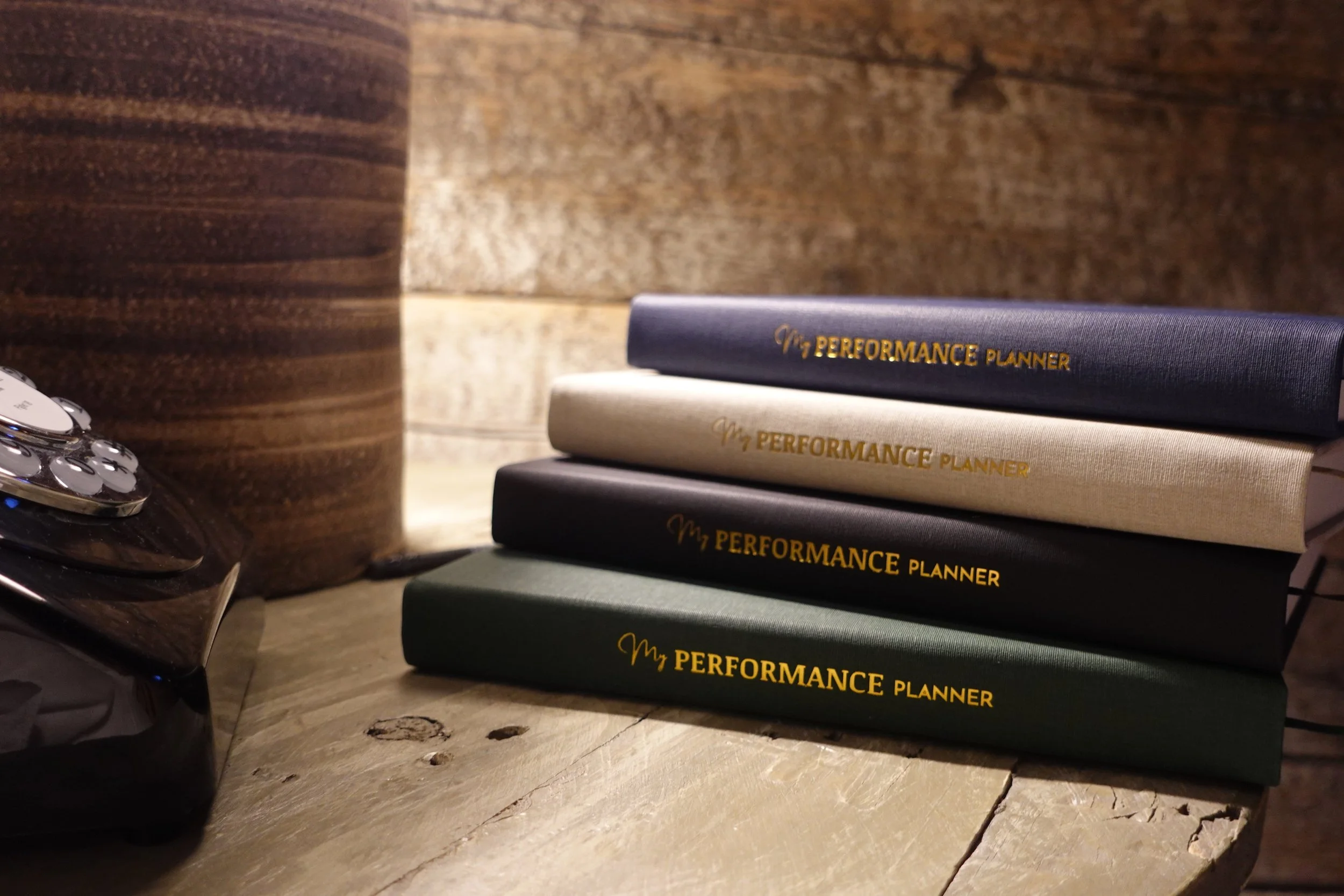 Stack of four black, white, and blue notebooks labeled "My Performance Planner" on a wooden surface with a wooden wall in the background.