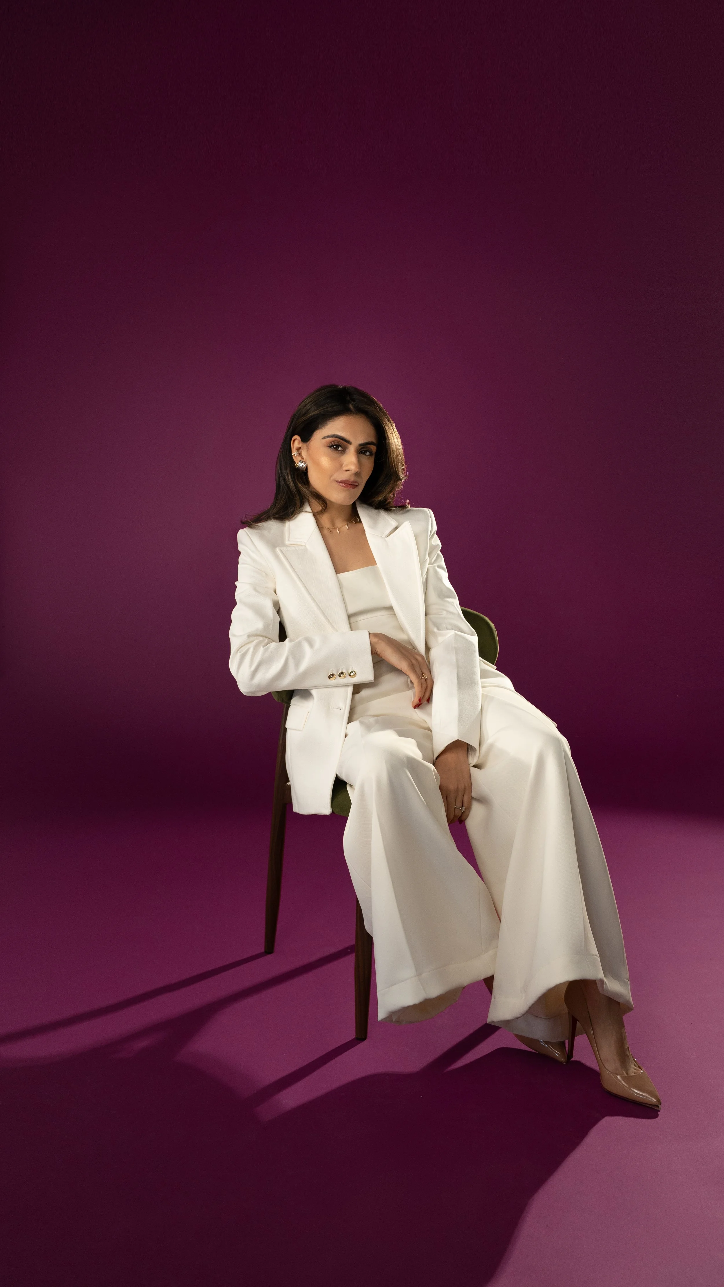 A woman wearing a white suit sitting on a chair against a purple background.