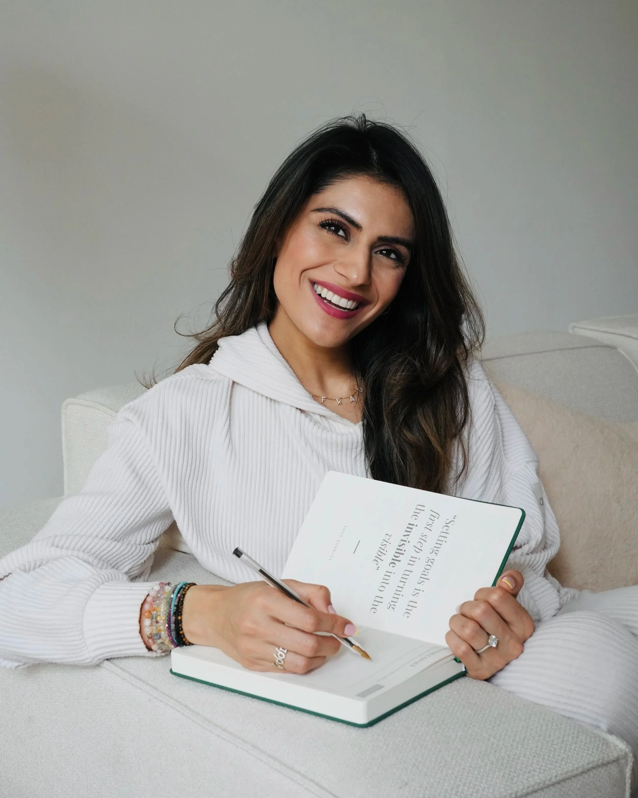 A woman with long dark hair, wearing a white sweater and accessories, is sitting on a white couch, smiling while writing in a book.