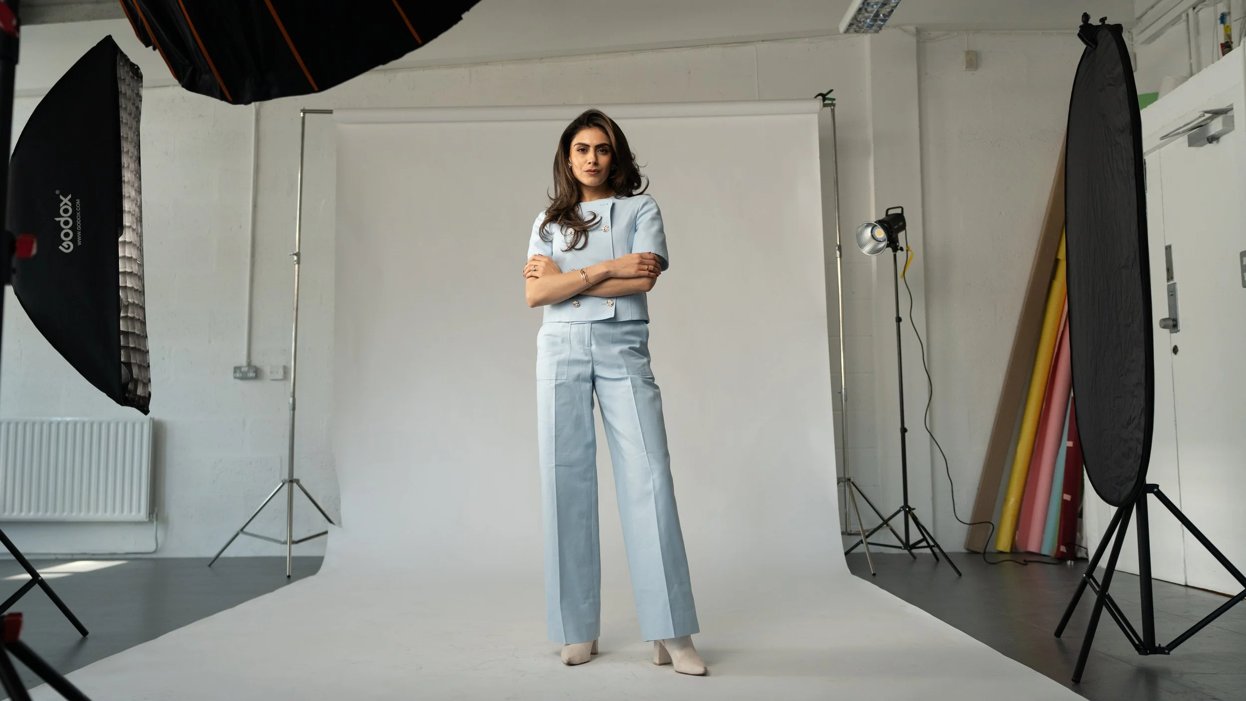 A woman with dark hair wearing a light blue pantsuit standing in a photography studio with studio lighting and a white backdrop.