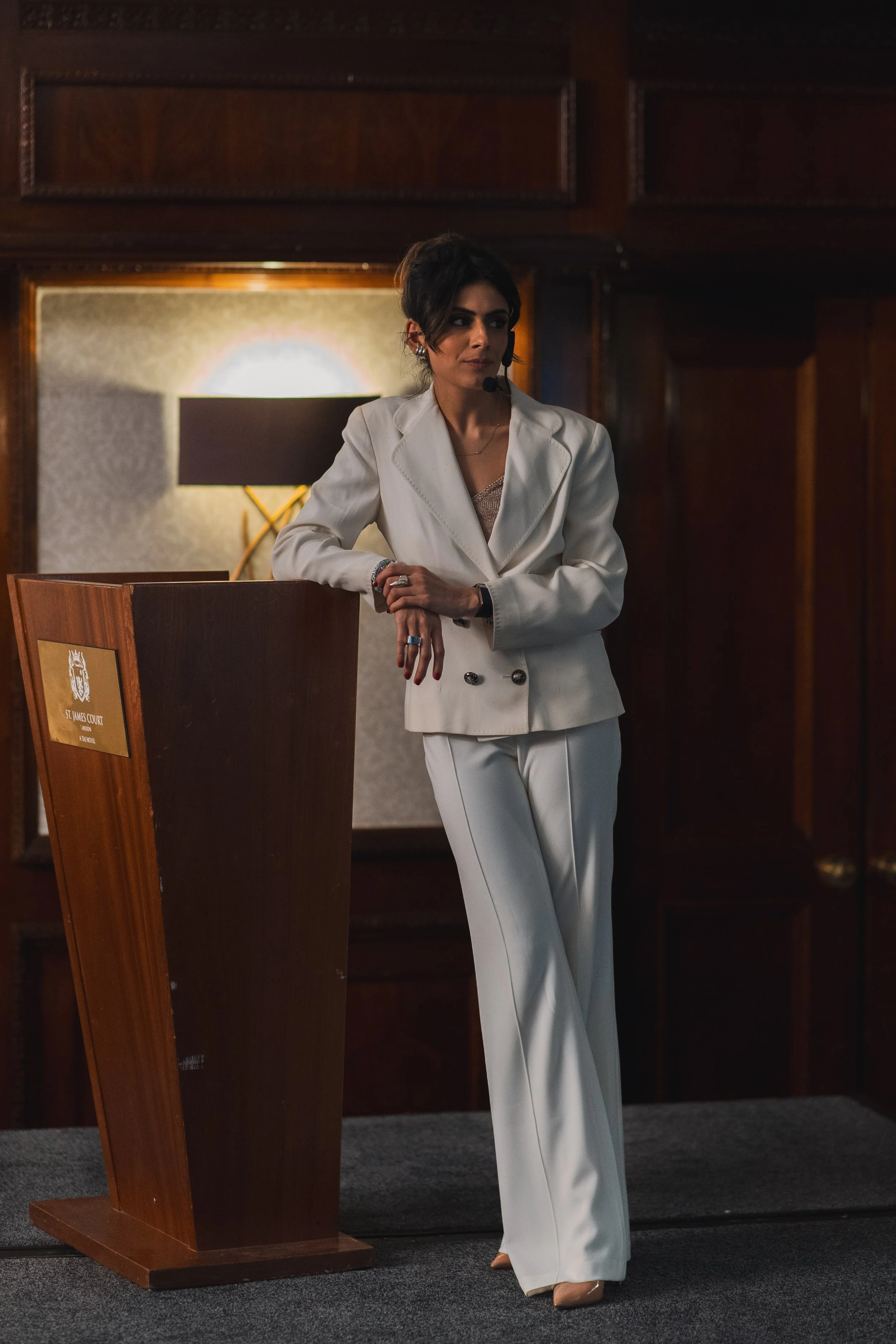 A woman wearing a white suit with wide-legged pants and a blazer, standing next to a wooden podium in a dimly lit room with dark wood paneling. She is wearing a headset microphone and has short dark hair, with her right hand resting on the podium and her left arm relaxed by her side.