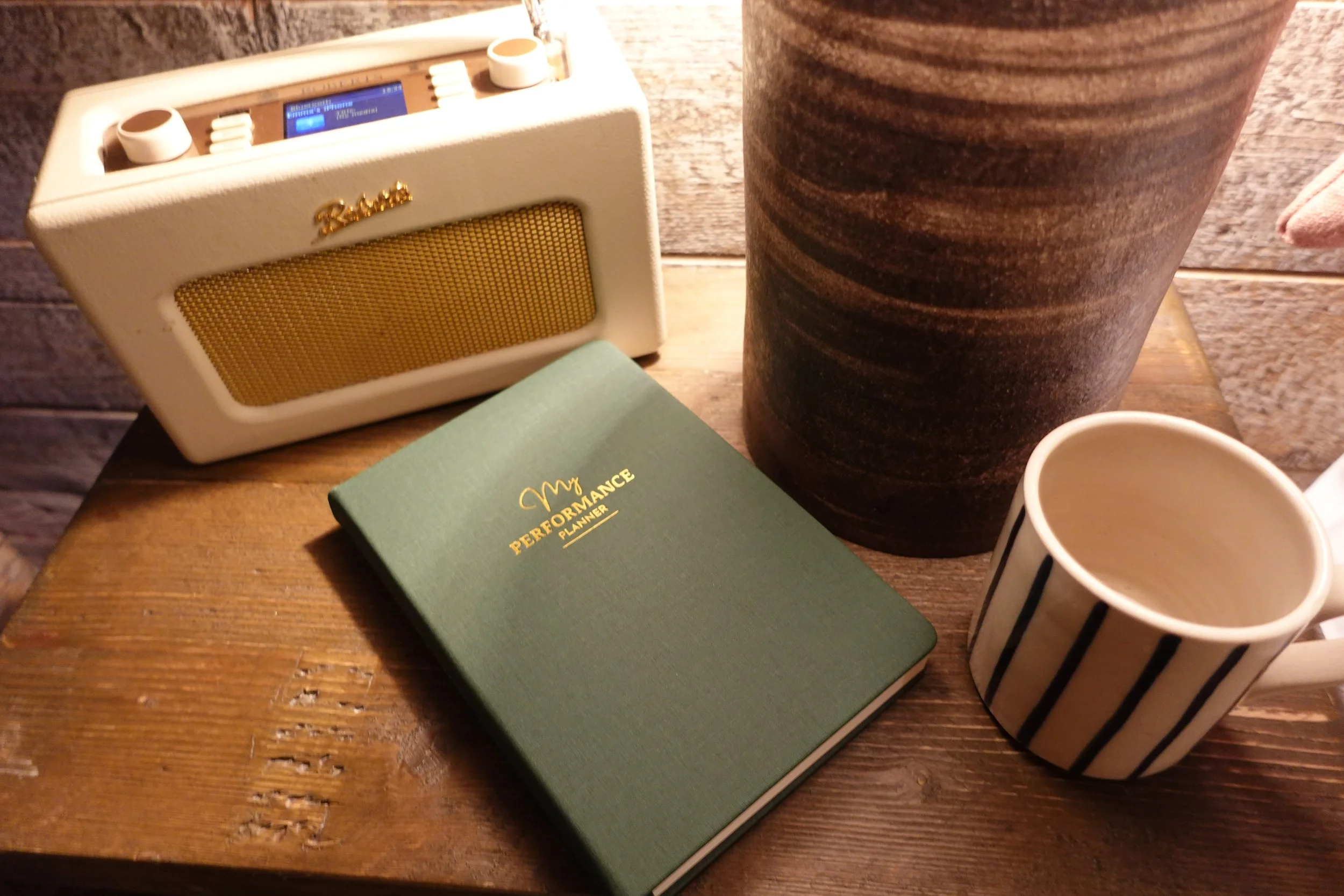 A wooden table with a small digital radio, a green planner titled "My Performance Planner," a large decorative brown and black vase, a striped paper cup, and a white cup, with a brick wall background.