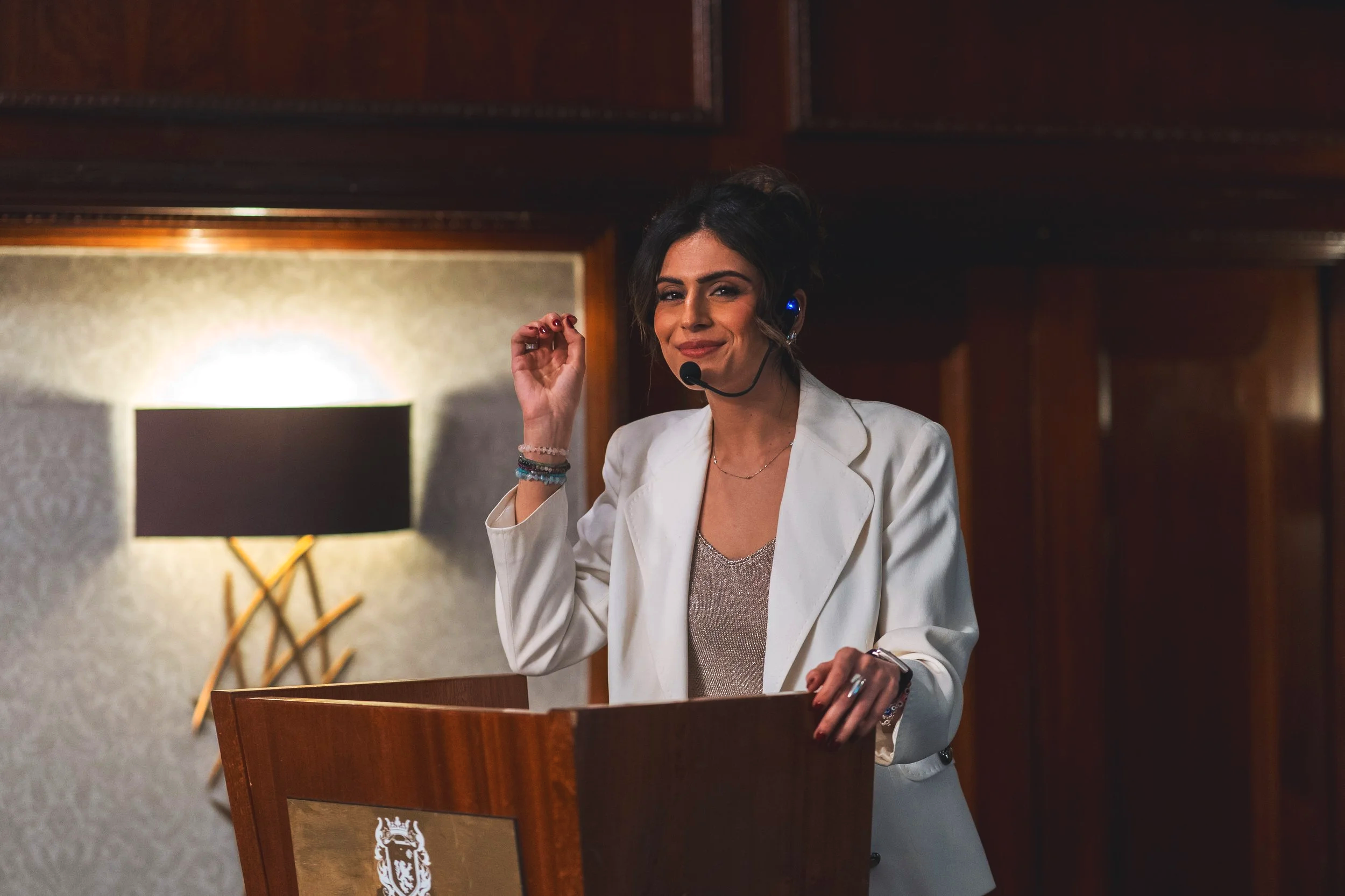 A woman with dark hair styled in loose waves, wearing a white blazer and a shiny top, standing at a wooden podium with a crest, smiling and adjusting her headset during a speech or presentation in a wood-paneled room.