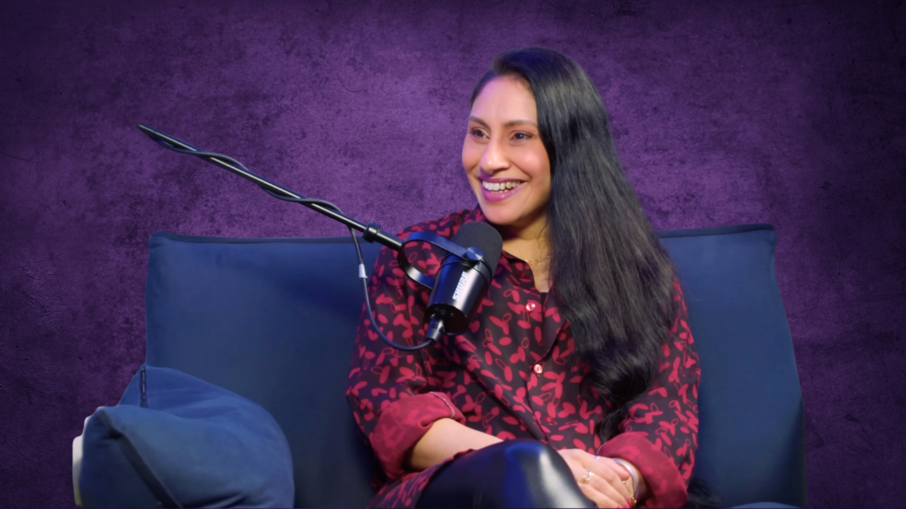A woman with long black hair smiling, sitting on a blue couch with a microphone in front of her against a purple textured background.
