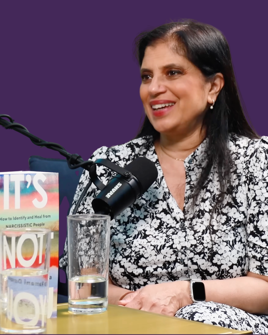 Smiling woman with dark hair wearing a black and white floral patterned shirt, sitting in front of a microphone, with a book titled 'It's Not You' and glasses of water on the table.
