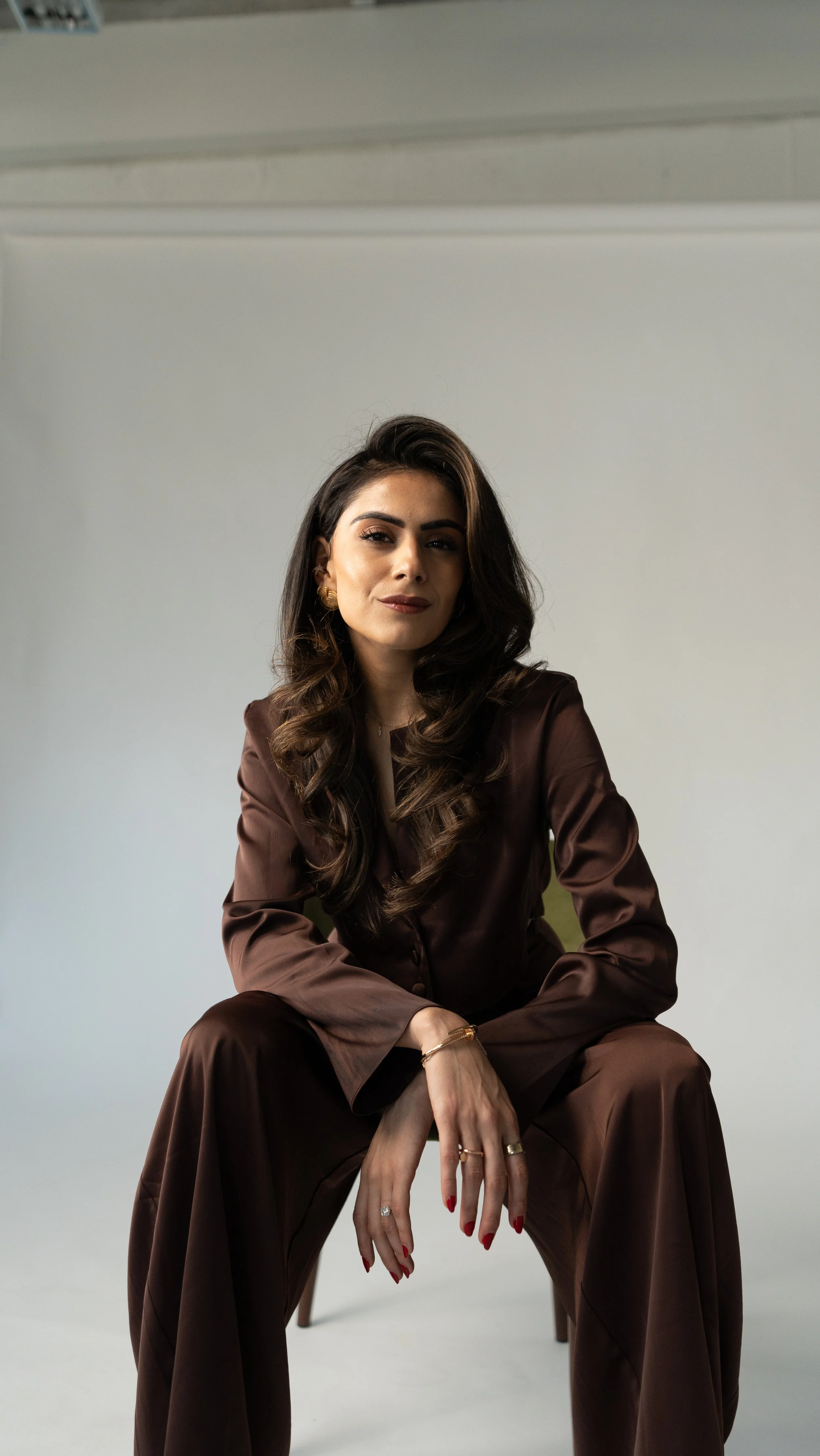 A woman with long dark wavy hair sitting on a chair against a plain background, wearing a brown satin blouse and matching wide-leg pants with gold jewelry and red nail polish.