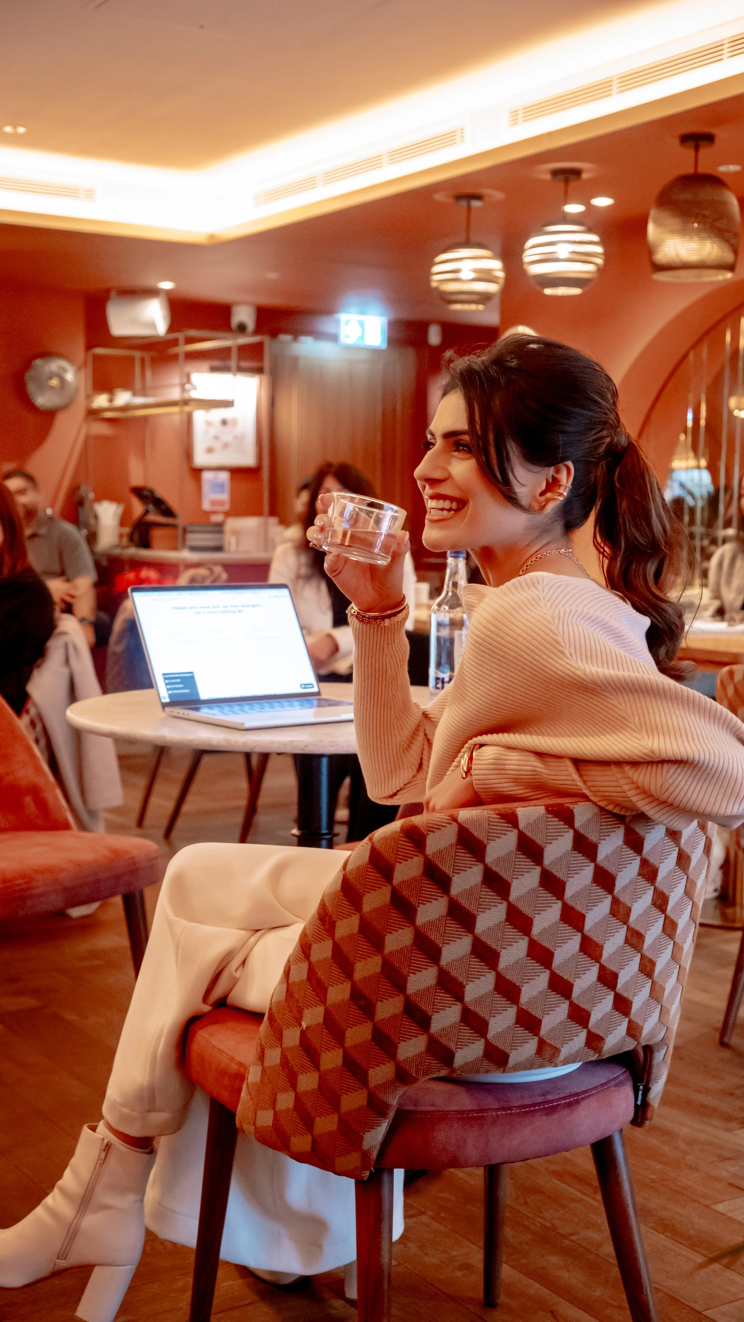Smiling woman with dark hair in a ponytail, sitting at a round table in a cozy restaurant, holding a glass of water. She is wearing a beige ribbed sweater and a patterned coat, with a laptop open in front of her. The background shows other diners, warm lighting, and wooden decor.