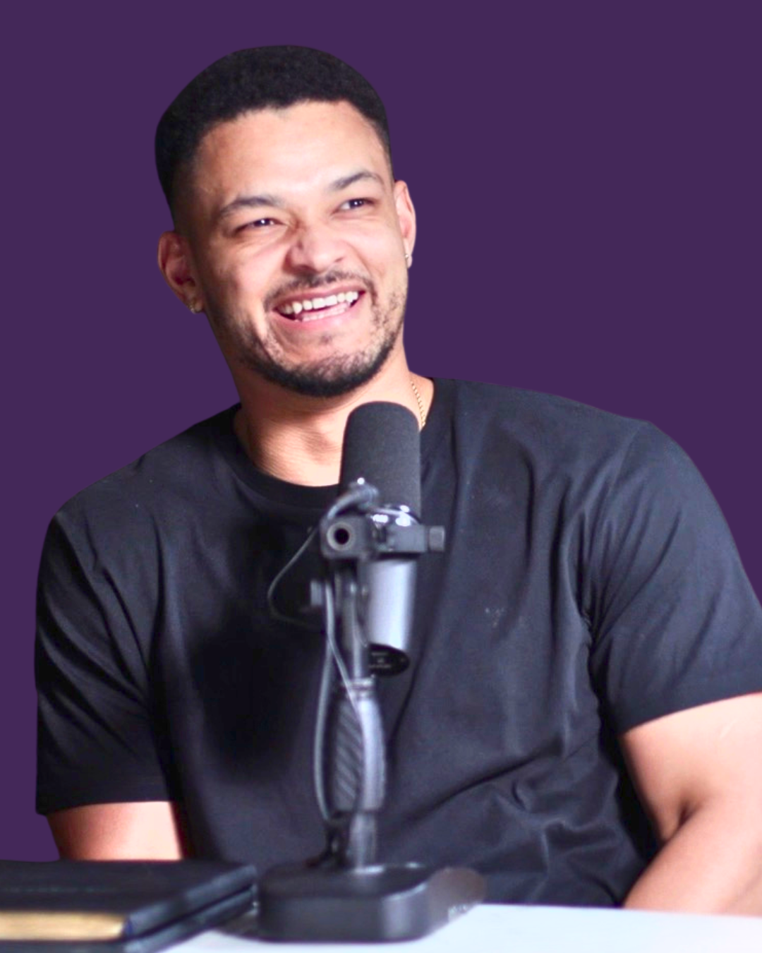 A young man with short dark hair and facial hair smiling while speaking into a microphone during a podcast or interview, sitting at a table with a purple background.