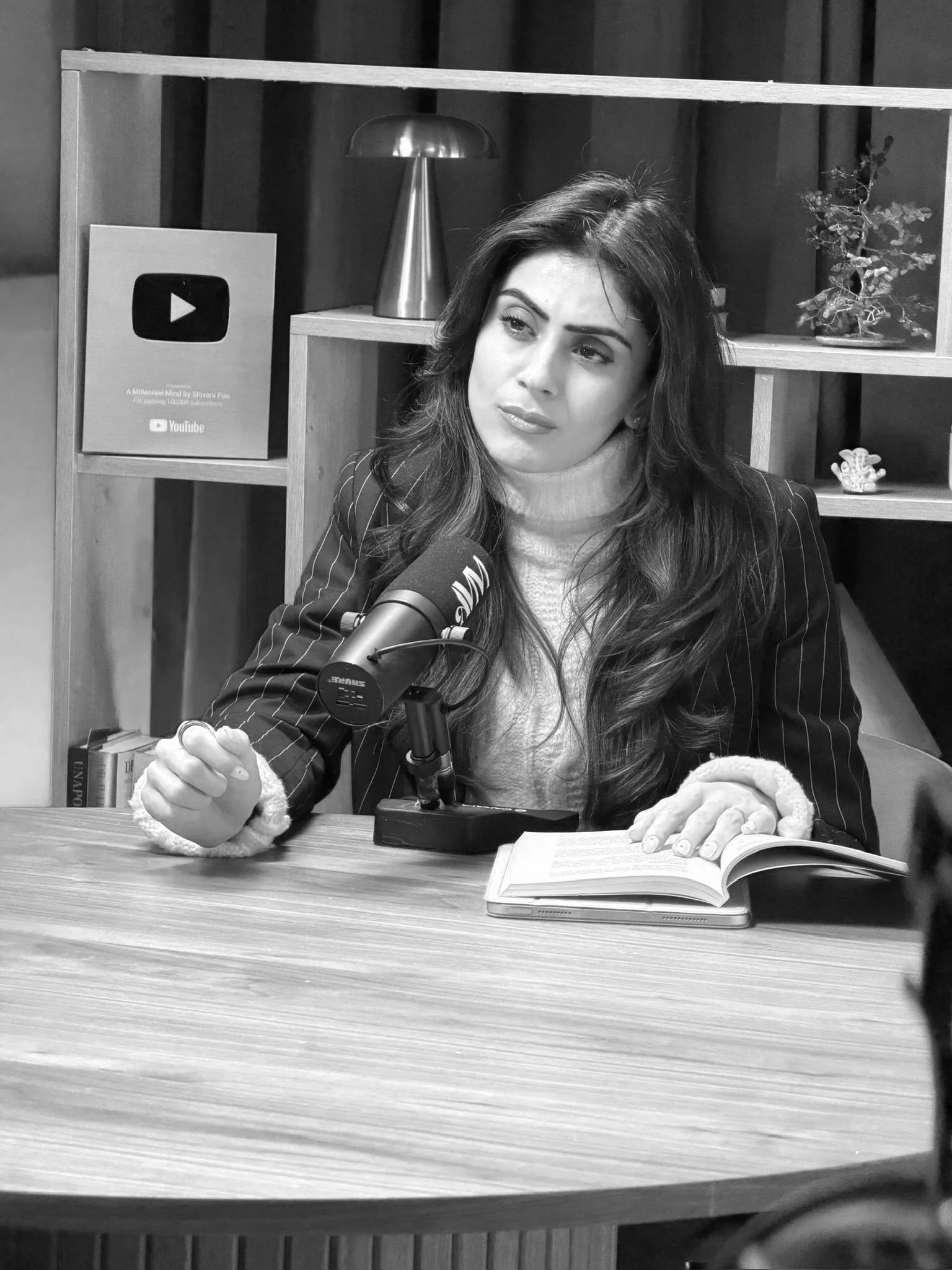 A woman with long dark hair, wearing a checkered blazer and white sweater, speaks into a microphone at a table with open books. Behind her is a bookshelf with a YouTube plaque, a lamp, and a potted plant, in a professional setting.