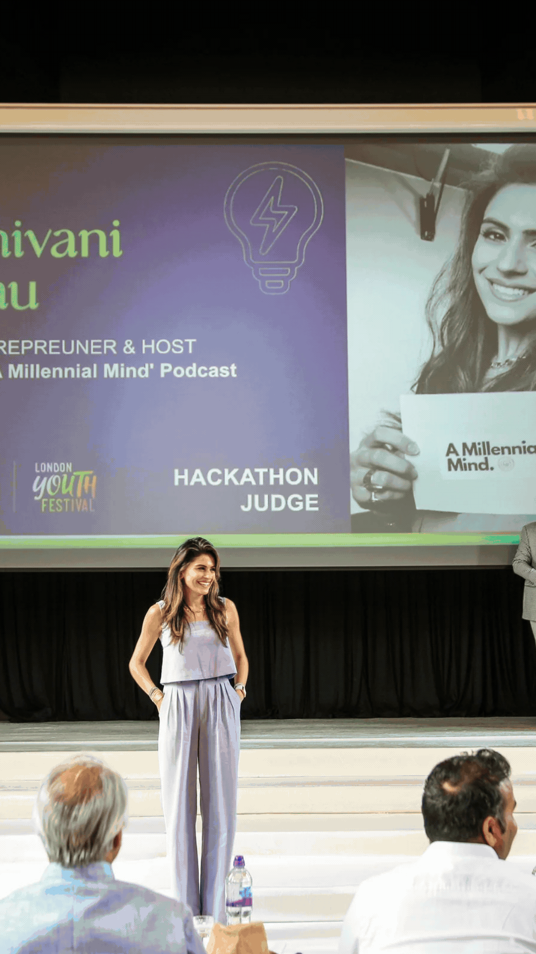 A woman stands on a stage smiling at an audience at a London youth festival event, with a large screen behind her displaying event information and her role as a hackathon judge.