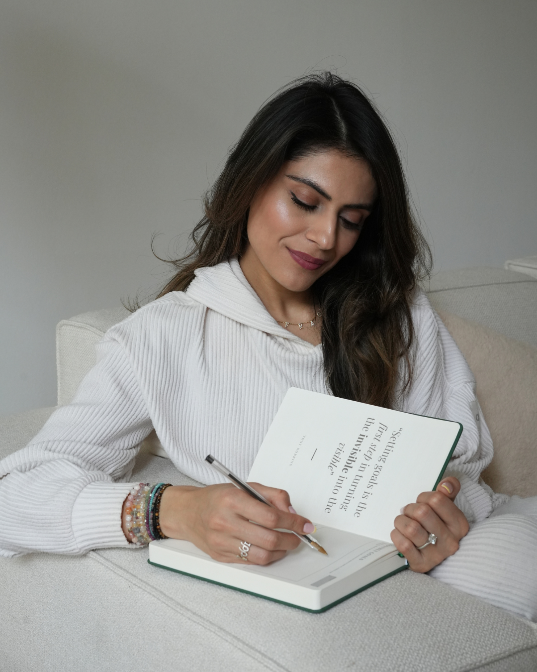 A woman with long dark hair, wearing a white sweater, sitting on a light-colored sofa, writing in a notebook with a pen.