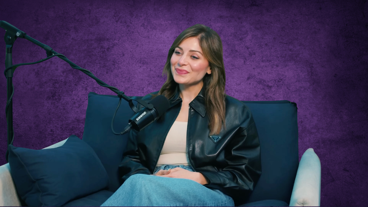A woman with long brown hair, dressed in a black leather jacket and beige top, sitting on a blue sofa with dark blue pillows, speaking into a professional microphone against a purple textured background.