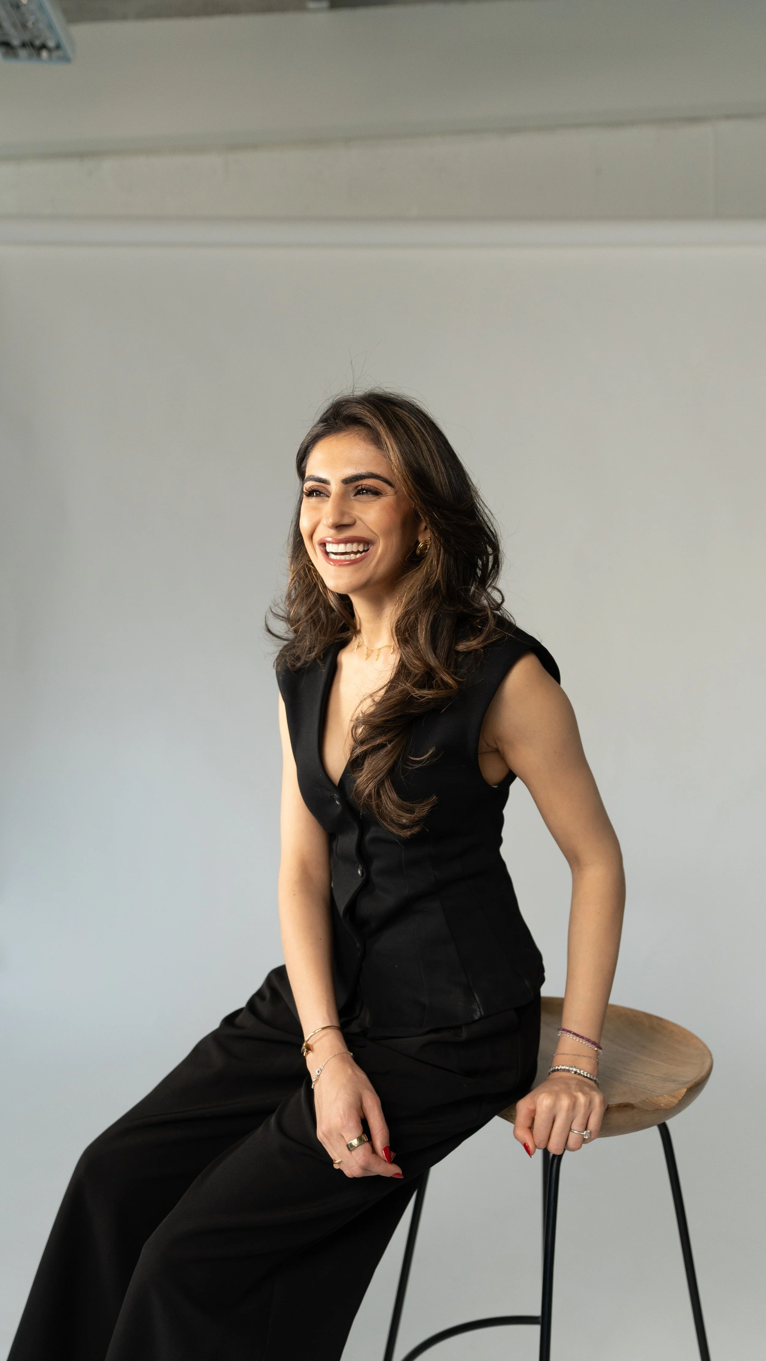 A woman with long wavy brown hair, dressed in a black sleeveless dress, sitting on a wooden stool with a light gray background.
