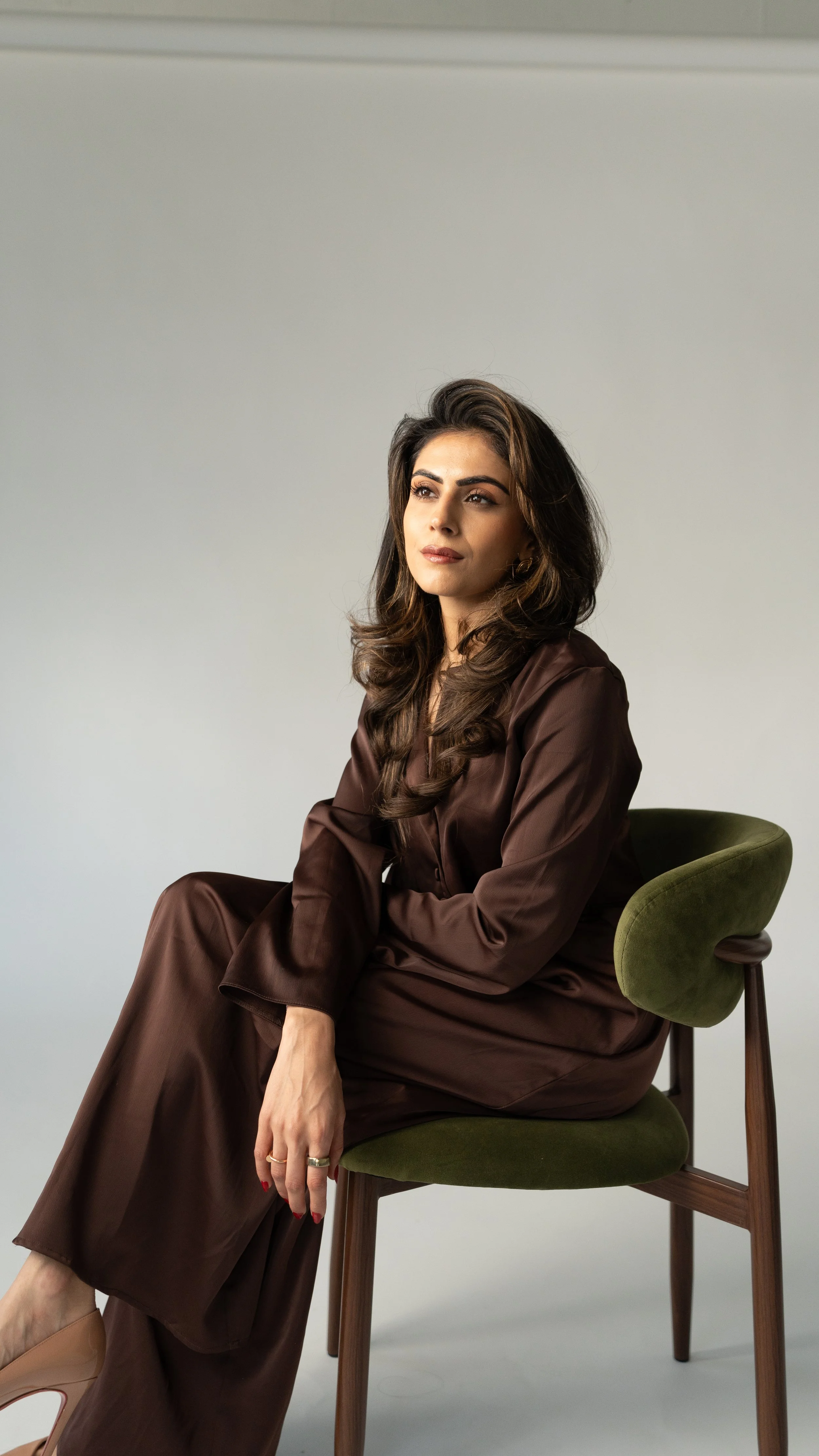A woman with long wavy brown hair, wearing a brown satin outfit, sitting on a green cushioned chair with wooden legs, against a plain light gray background.
