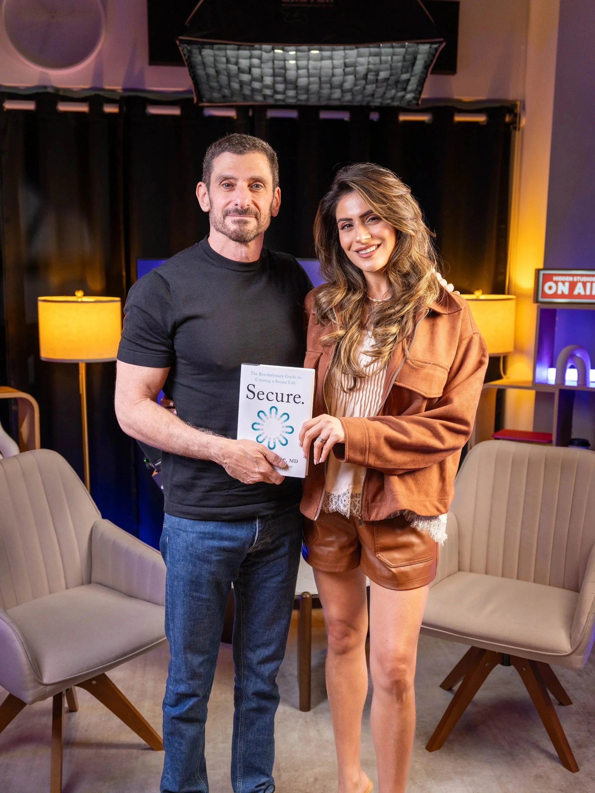 A man and a woman standing close together in a room, smiling, with chairs and lamps in the background. The man holds a book titled 'Secure'. The woman is winking and wearing a brown jacket and shorts, while the man is wearing a black t-shirt and jeans.