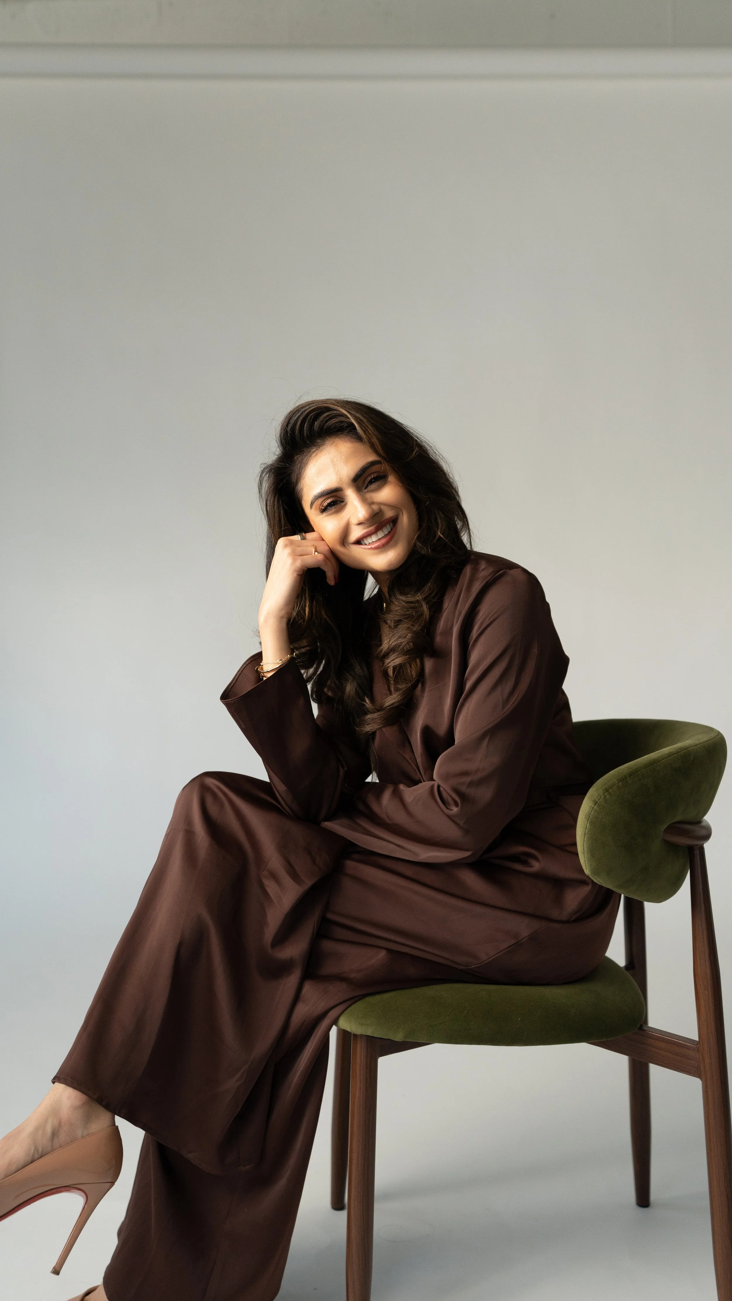 A woman with dark hair, smiling, sitting on a green chair, wearing a brown satin suit and beige high heels, against a plain gray background.
