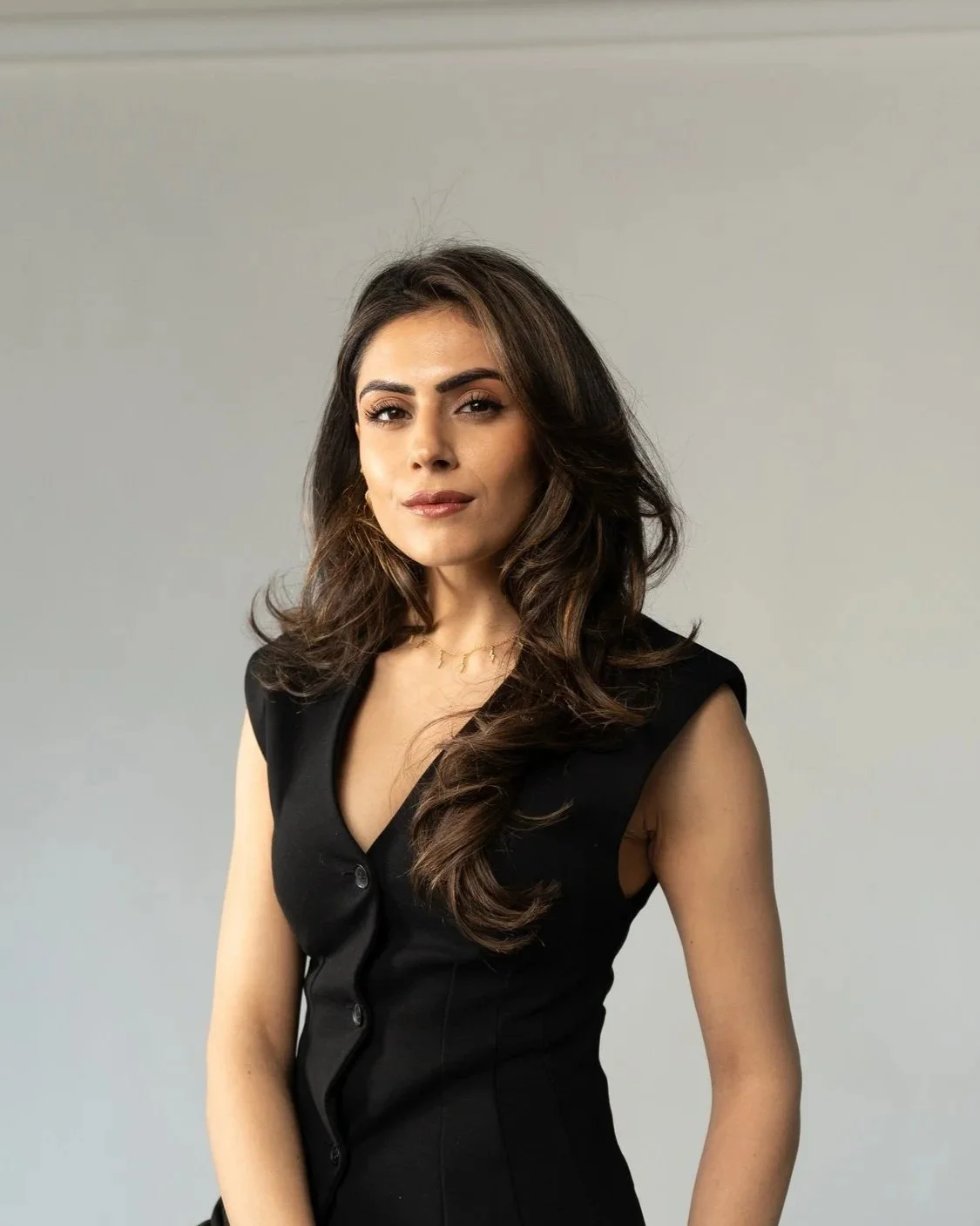A woman with long wavy brown hair wearing a sleeveless black dress and a delicate necklace stands against a plain gray background.
