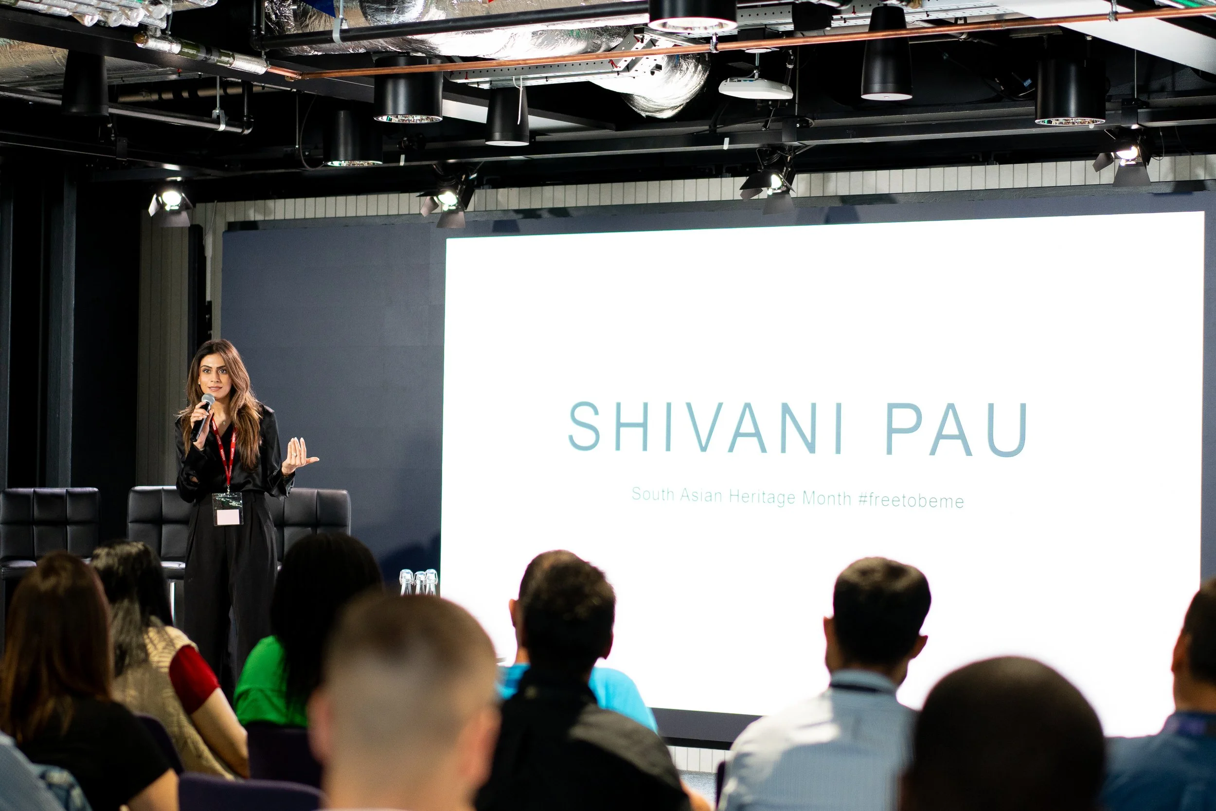 A woman with long hair speaking into a microphone on stage at event about South Asian Heritage Month, with large screen displaying her name 'Shivani Pau' and audience capturing her presentation.